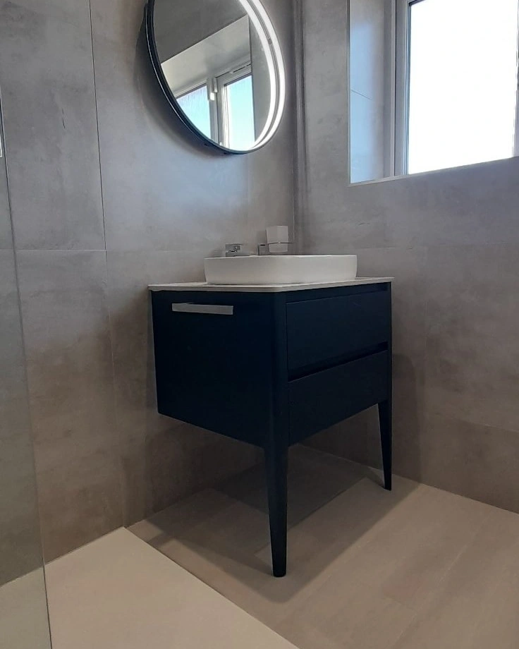 Modern bathroom corner with a wall-mounted dark blue cabinet, white vessel sink, round mirror, and window.