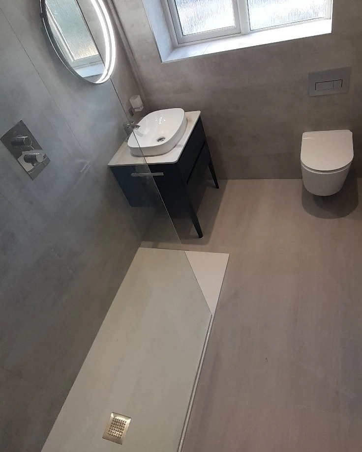Minimalist bathroom with walk-in shower, black vanity with white vessel sink, round illuminated mirror, and wall-mounted toilet under frosted window.