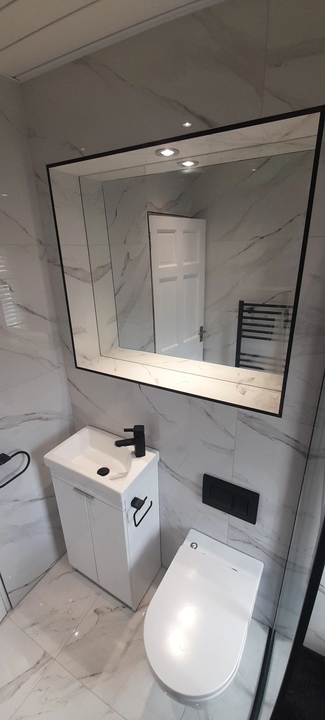 Modern bathroom with white marble tiles, a white toilet, a small white sink with a black faucet, and a large rectangular mirror above.