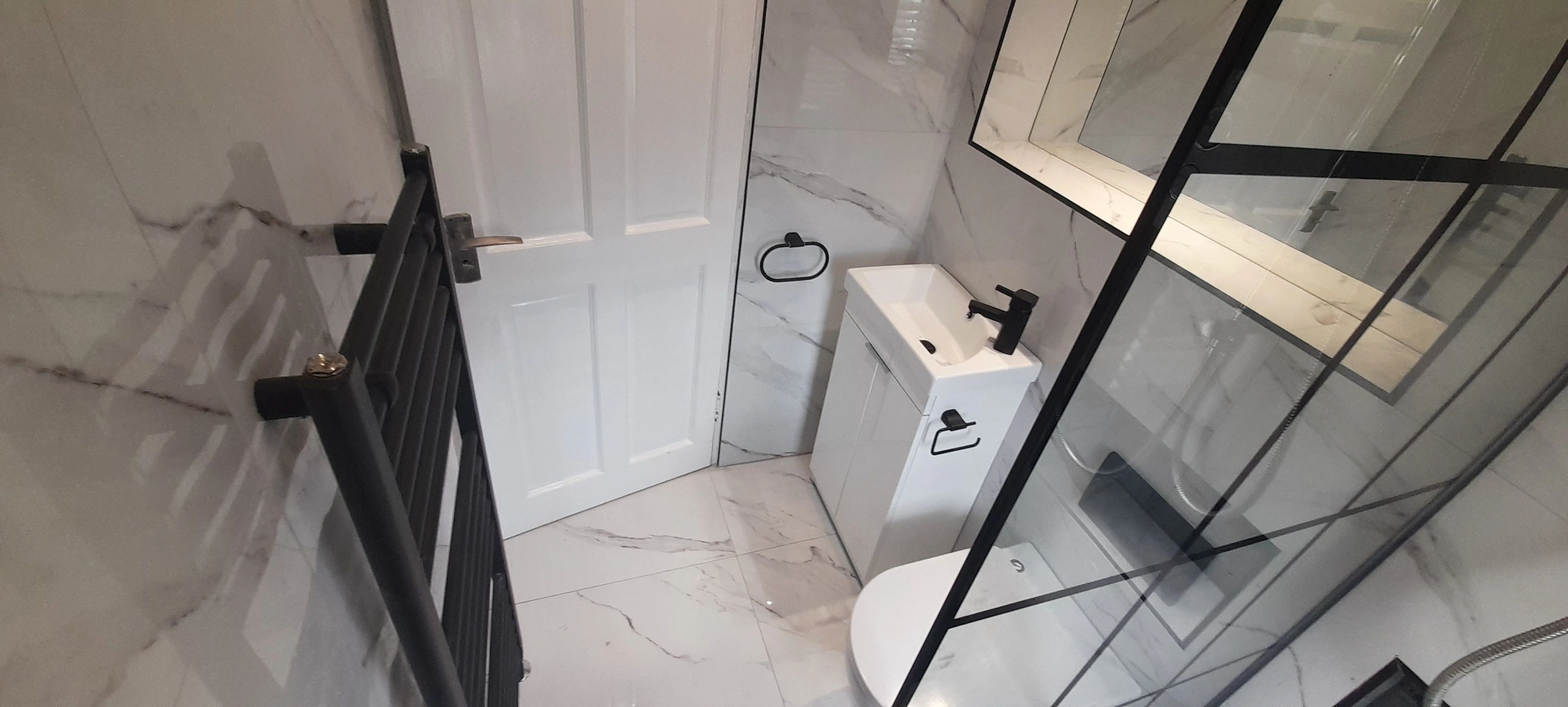 Small modern bathroom with white marble floor and walls, black-framed glass shower, white door, white sink with black faucet, and black heated towel rail.