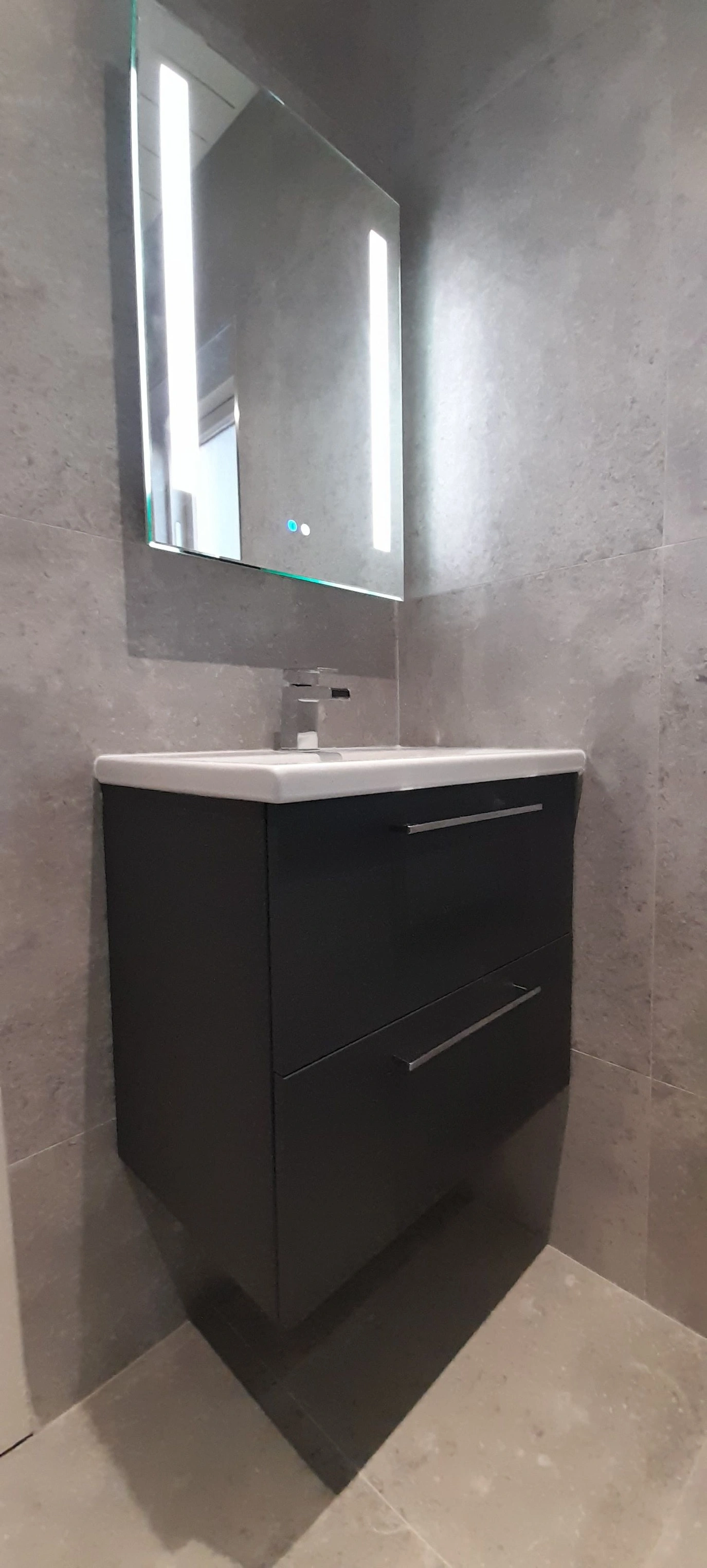 Modern bathroom vanity with black cabinet, white sink, and lit rectangular mirror on gray tiled wall.