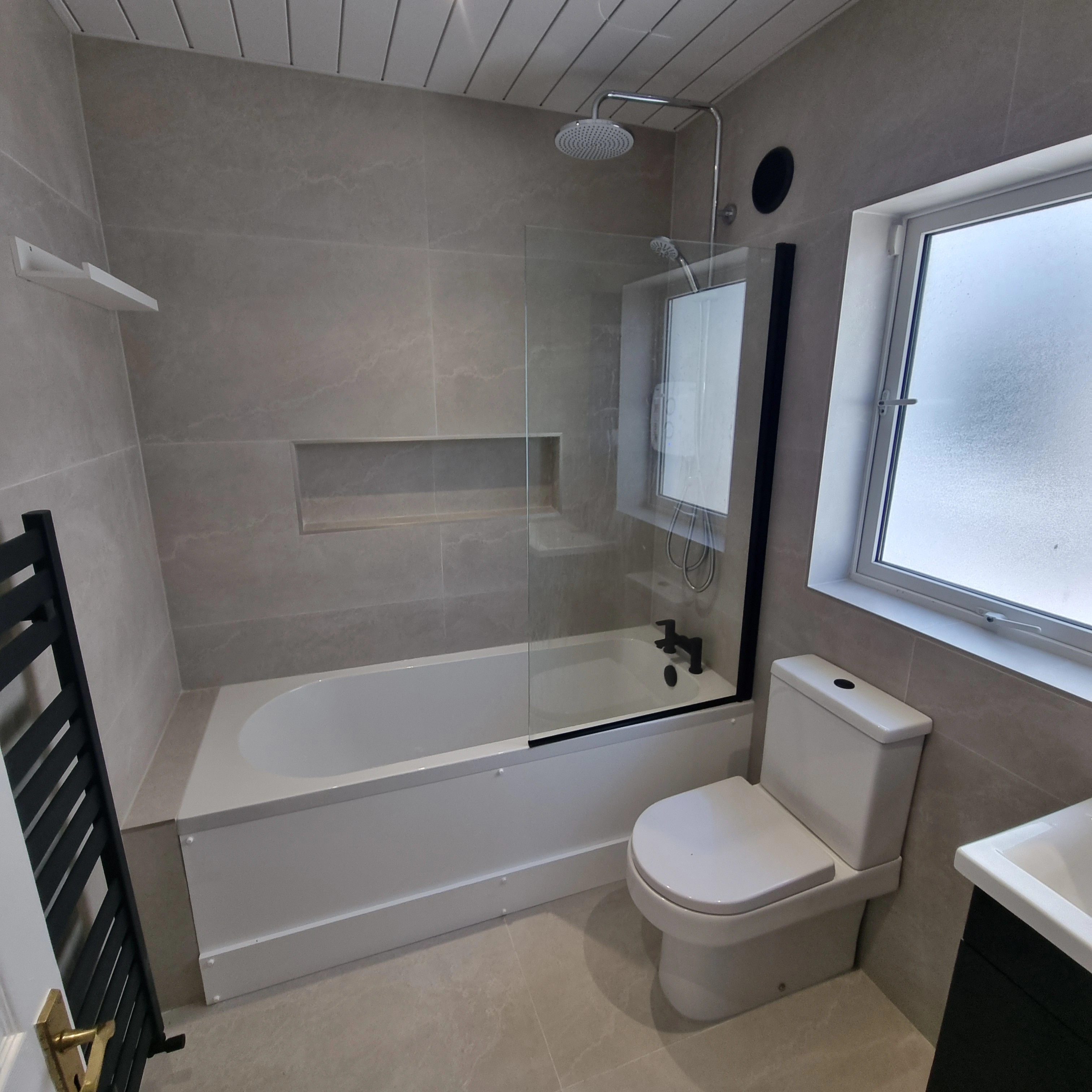 Modern bathroom with white bathtub and black fixtures, a frosted window, white toilet, and light gray tiled walls and floor.