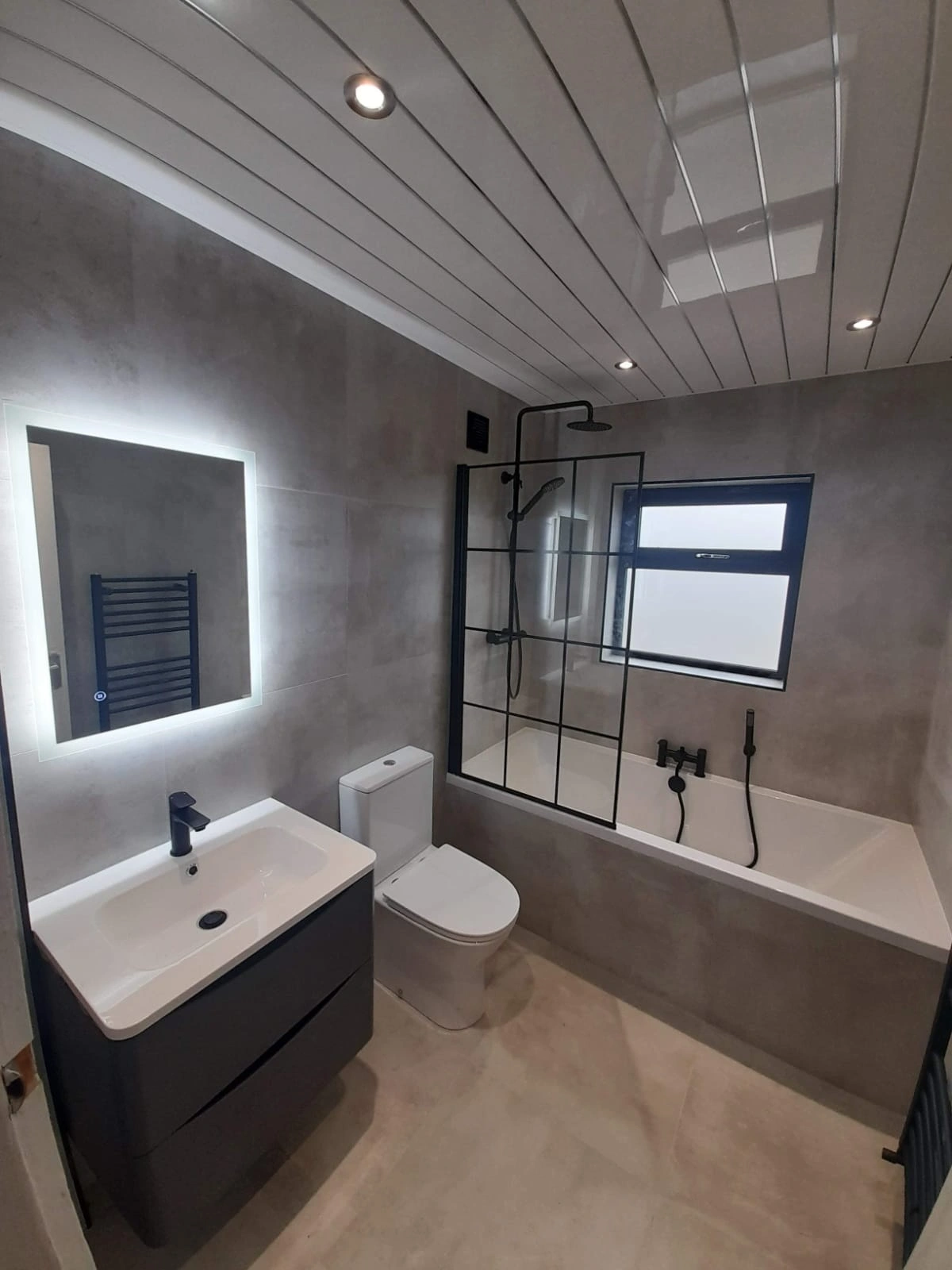 Modern bathroom with gray tile walls, a white toilet, a vanity with sink and illuminated mirror, and a bathtub with black-framed glass shower screen and matte black fixtures.