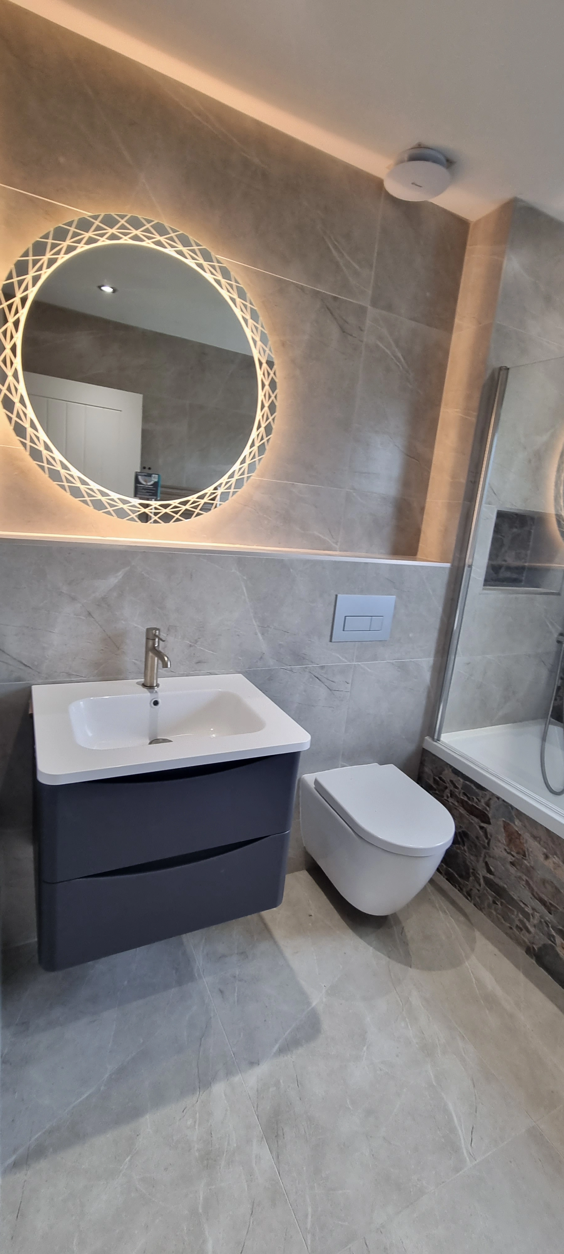Modern bathroom with a wall-mounted sink and cabinet, illuminated round mirror, wall-hung toilet, and bathtub with glass screen and stone trim.