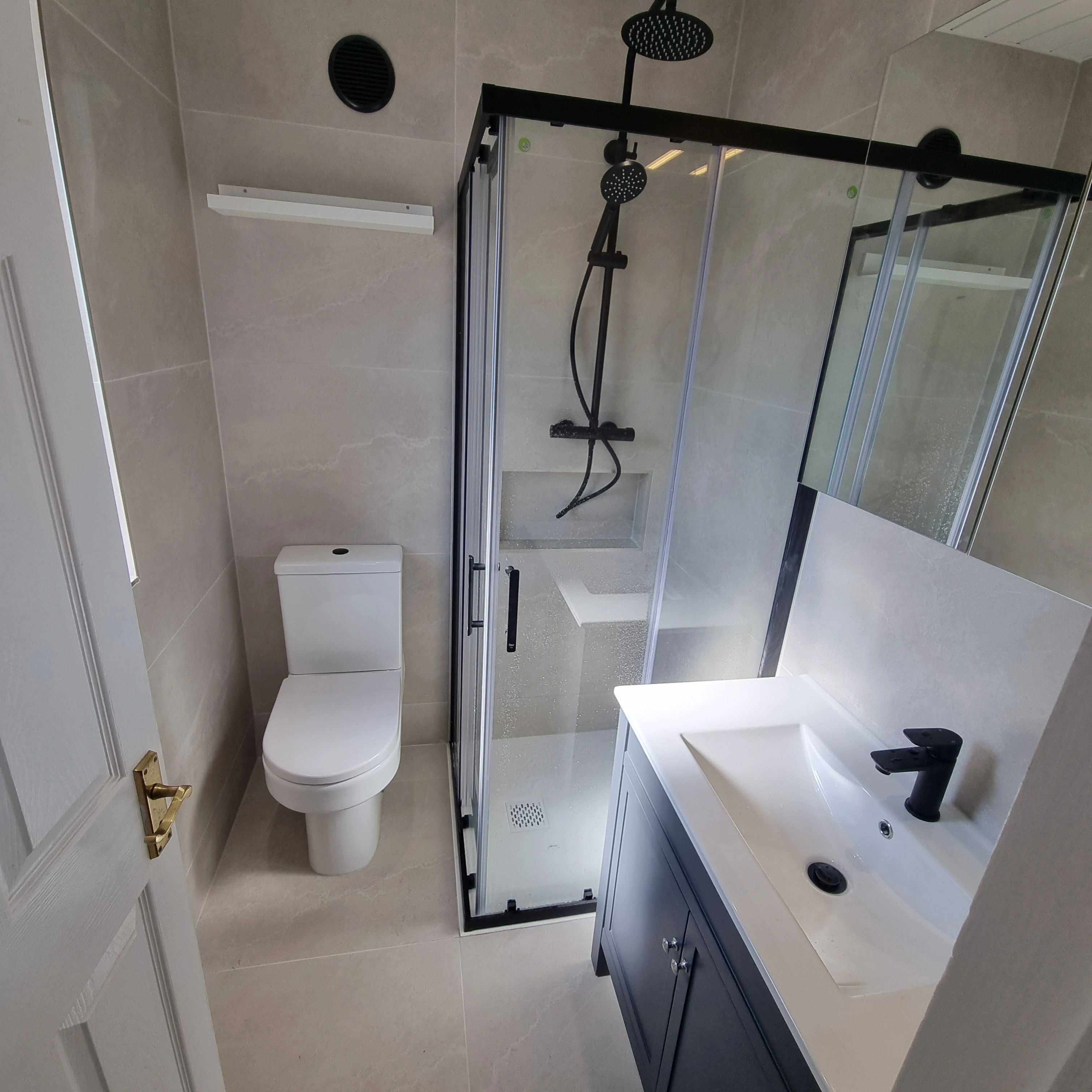 Modern bathroom with a white toilet, glass-enclosed shower with black fixtures, and a white sink with black faucet on a dark vanity.