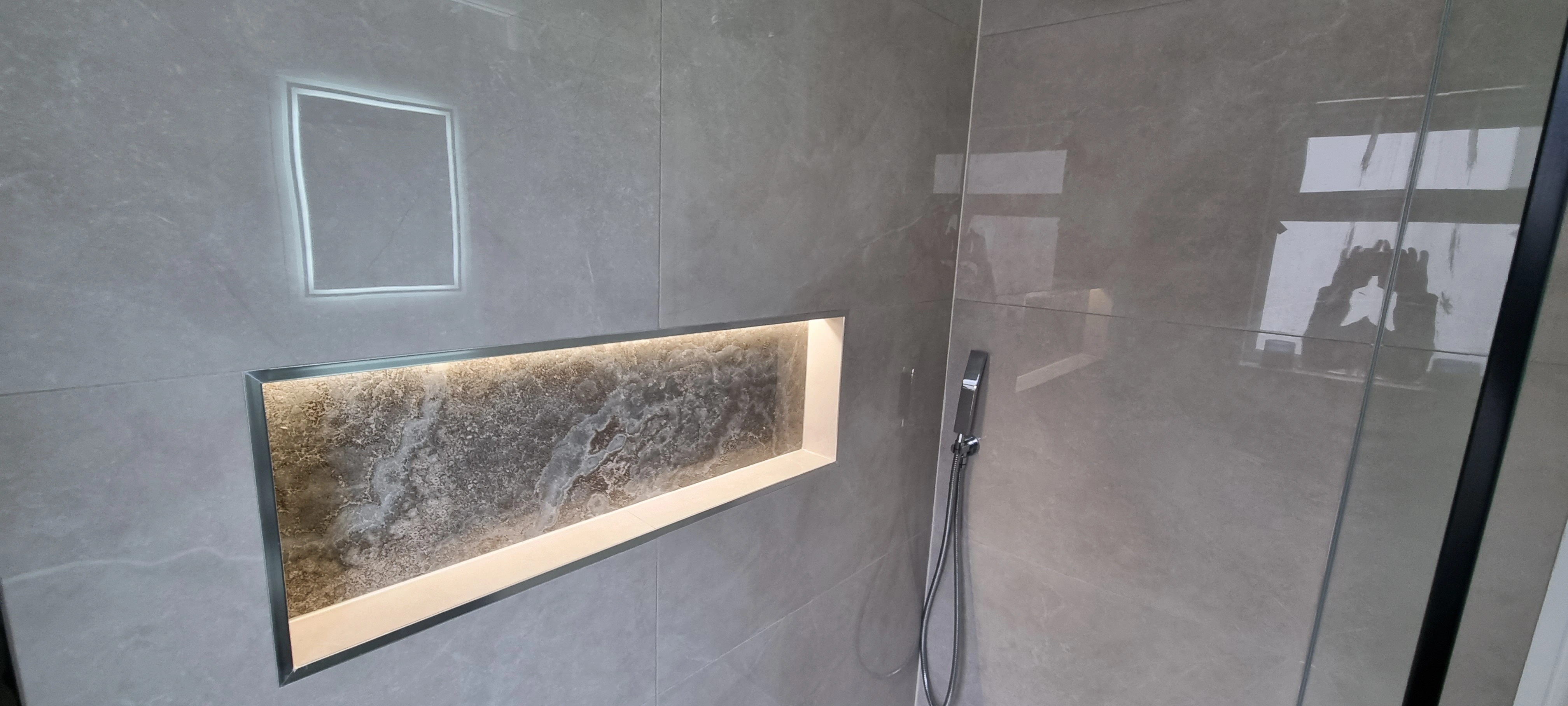 Modern shower area with gray tiled walls, built-in rectangular niche with textured stone and lighting, and a handheld showerhead.
