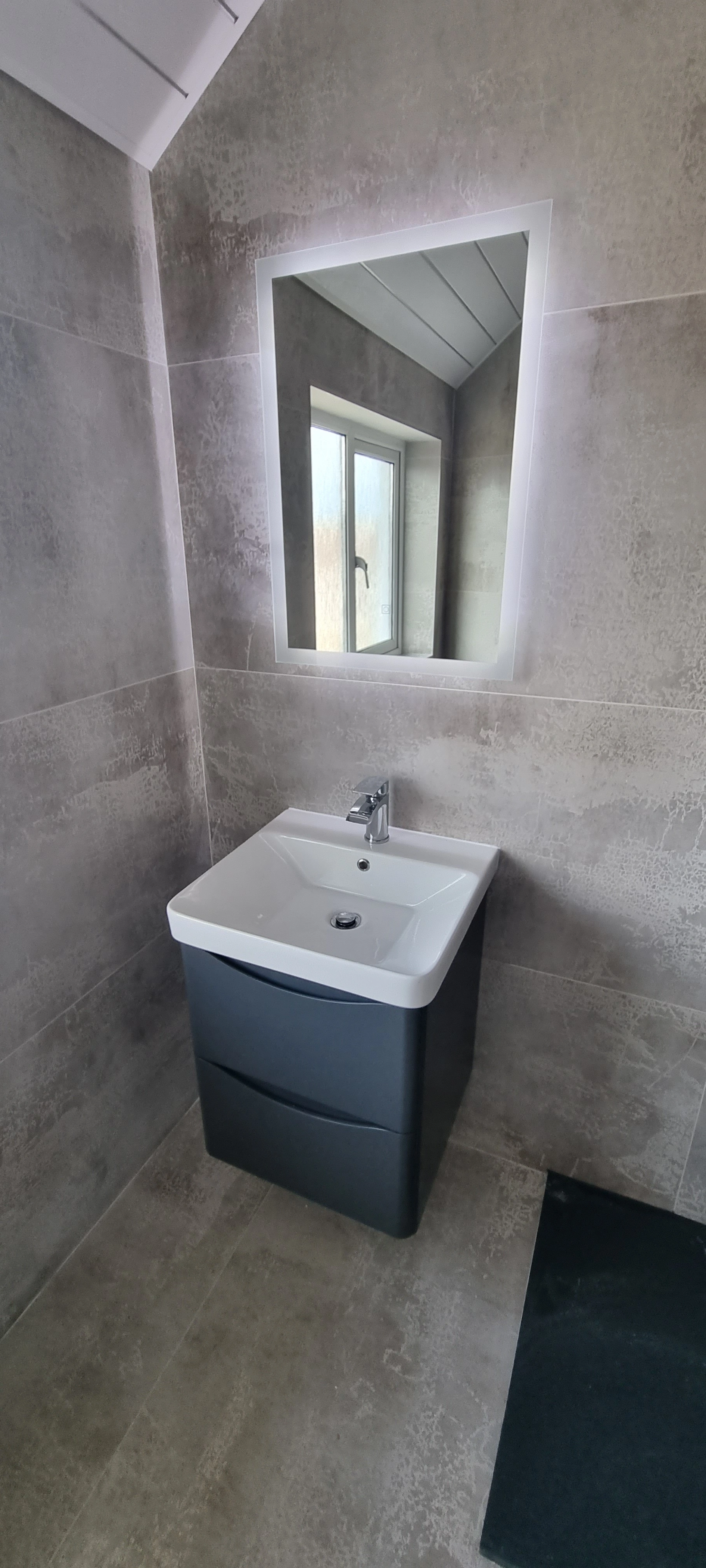 Modern bathroom corner with grey tiled walls and floor, a white sink with dark vanity cabinet, and a rectangular backlit mirror reflecting a window.
