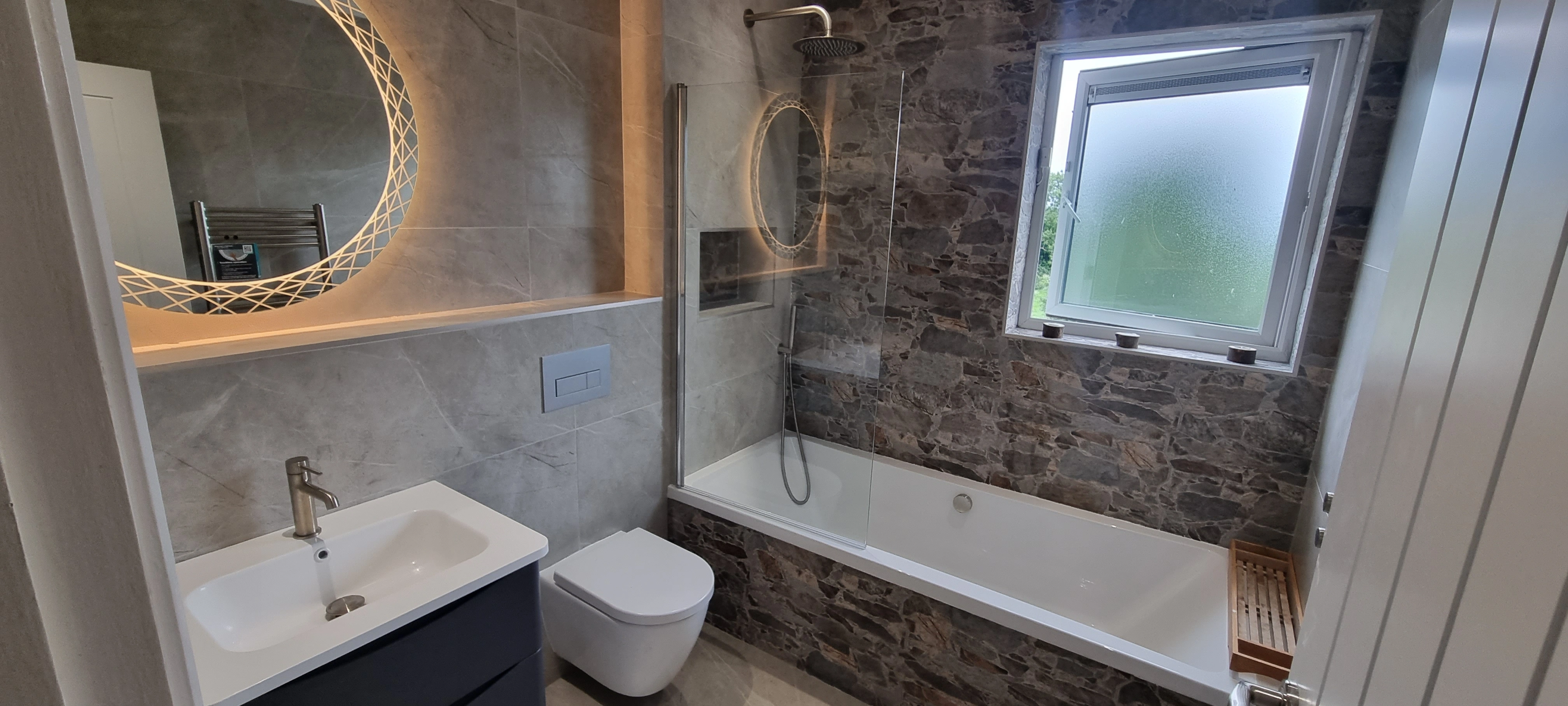 Modern bathroom with a wall-mounted toilet, a white sink with vanity, a bathtub with stone tile surround, and a round illuminated mirror.
