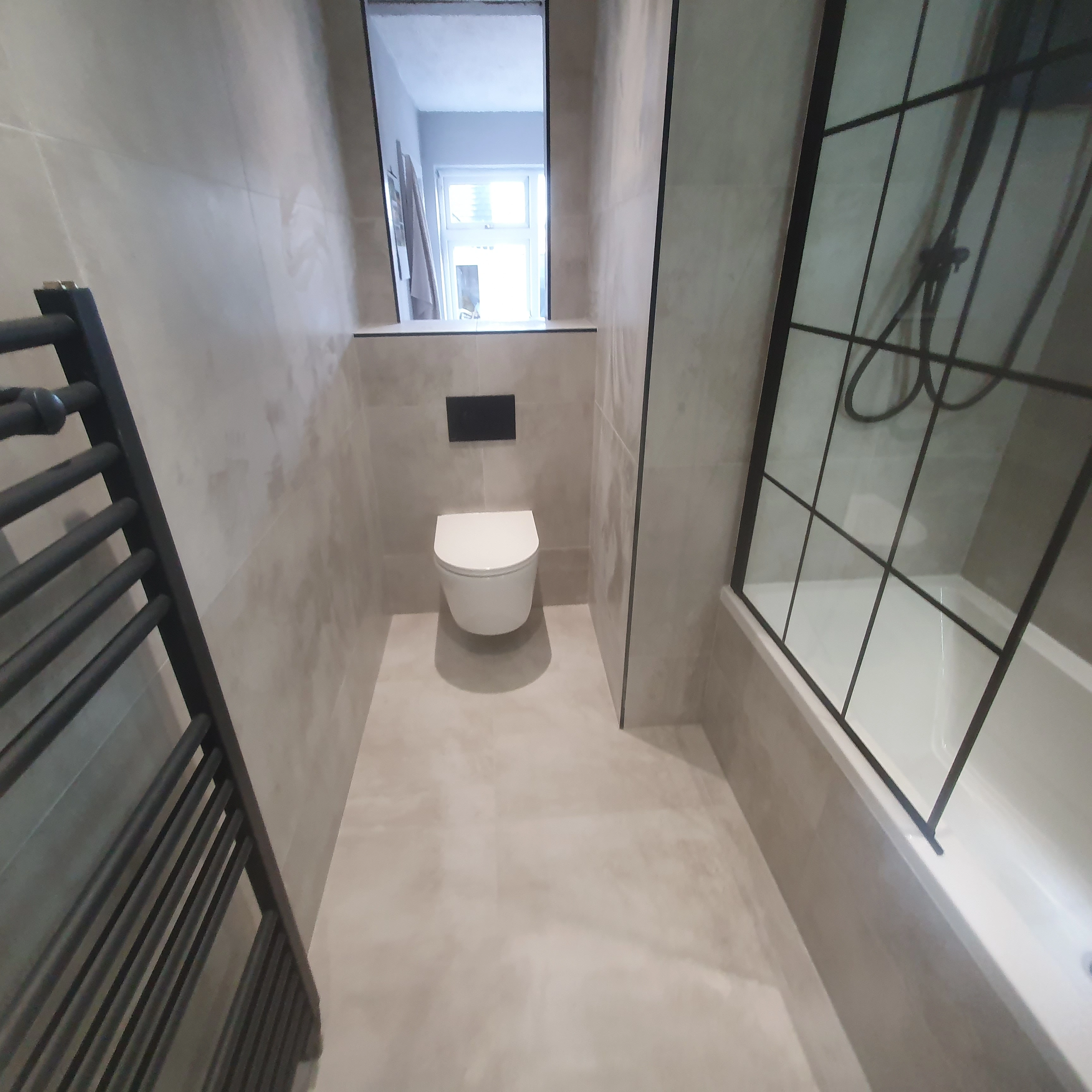 Modern bathroom with wall-mounted toilet, gray tiled walls and floor, black towel radiator, and bathtub with glass panel and black grid frame.