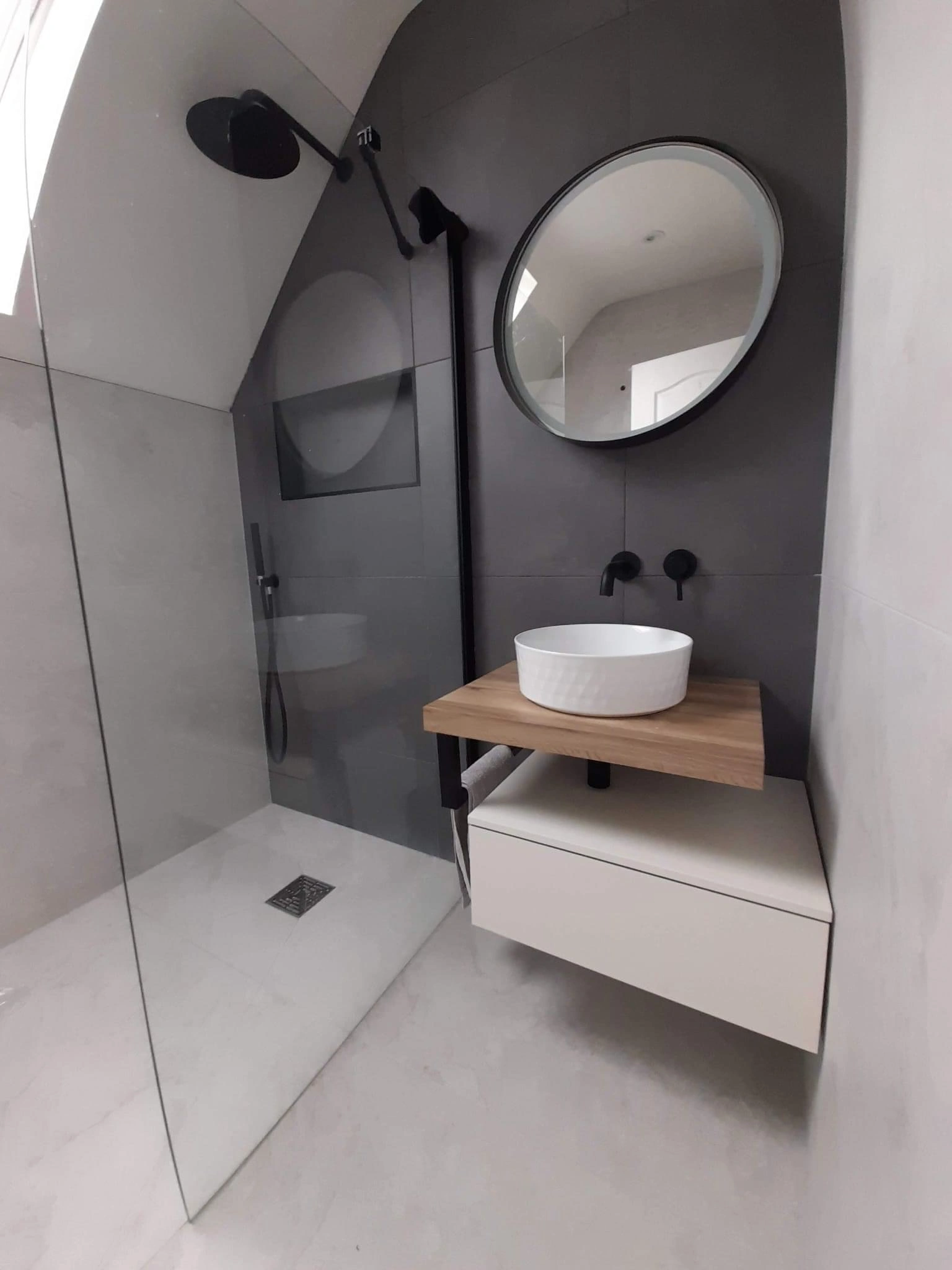 Modern bathroom with a round mirror, white vessel sink on a wooden shelf, black wall-mounted faucets, and a glass shower enclosure with a black showerhead.