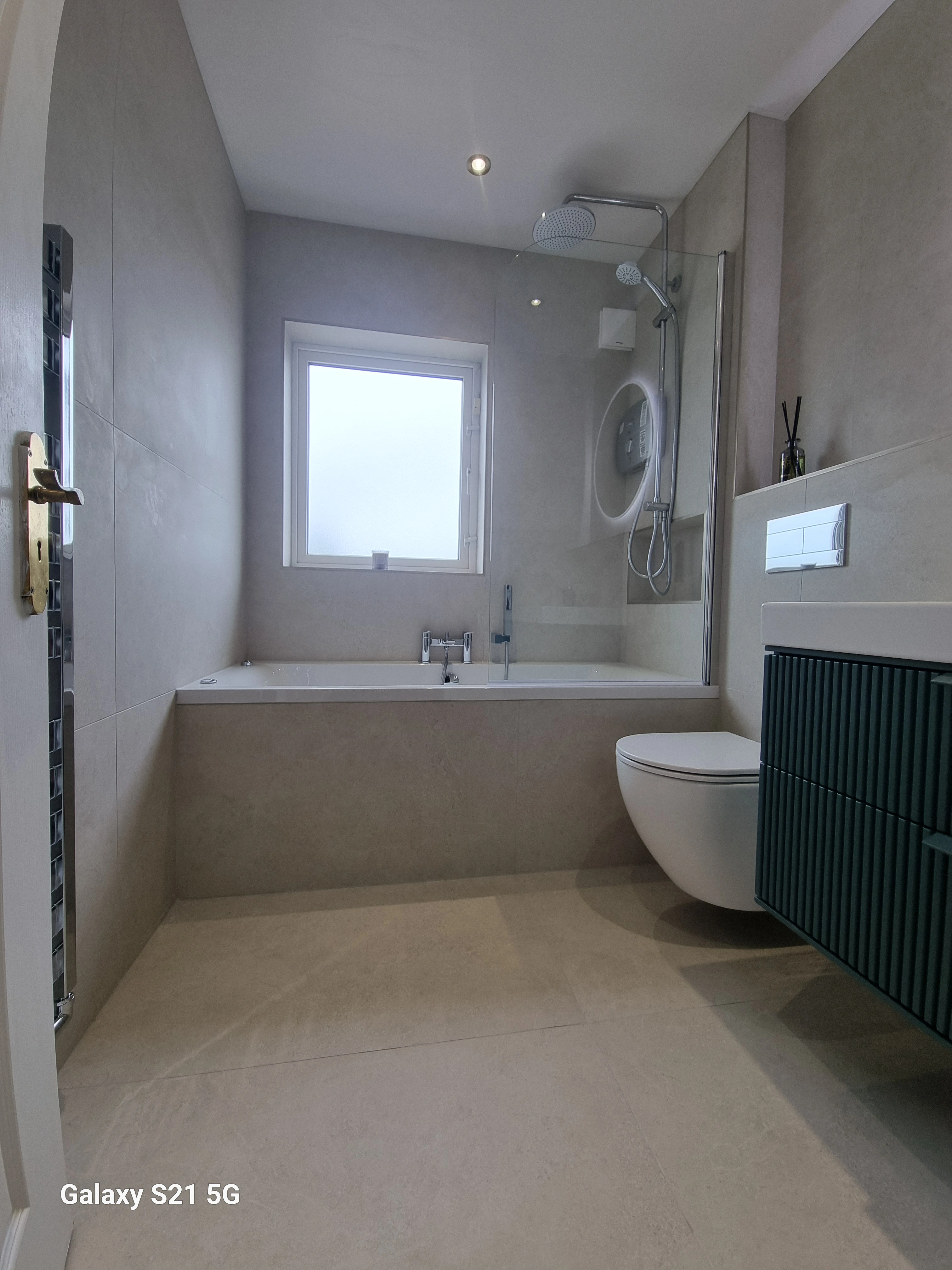 Modern bathroom with beige tiled walls and floor, featuring a bathtub under a frosted window, a wall-mounted toilet, a shower head with glass partition, and a green bathroom vanity.