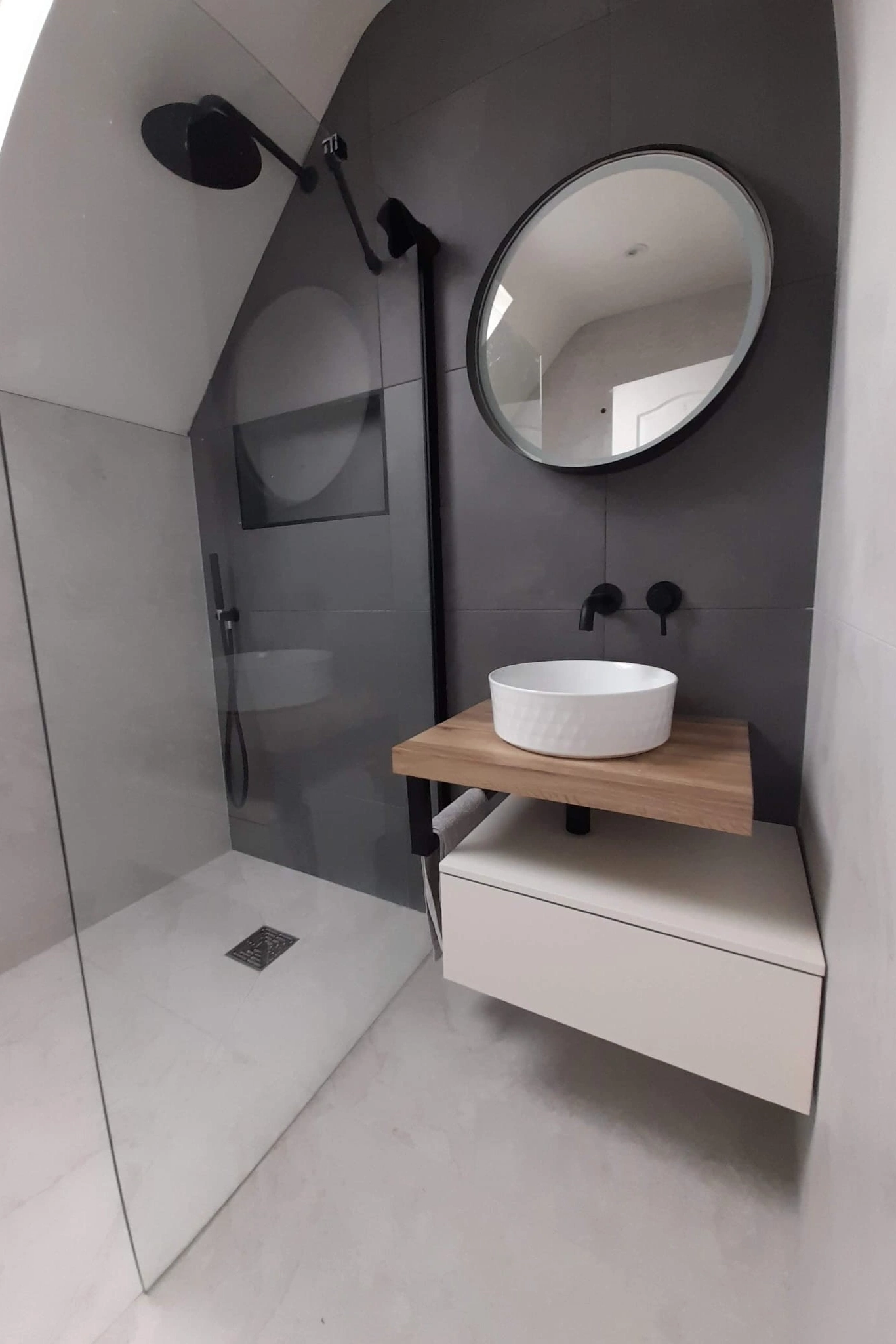 Modern sleek bathroom with a walk-in shower featuring a black showerhead and a floating wood and white vanity with a round vessel sink and circular mirror.
