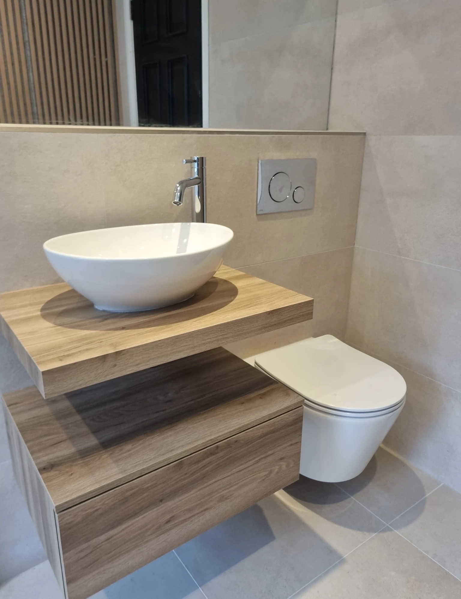 Modern bathroom with wall-mounted white toilet and a wooden floating vanity holding an oval white vessel sink and a chrome faucet.