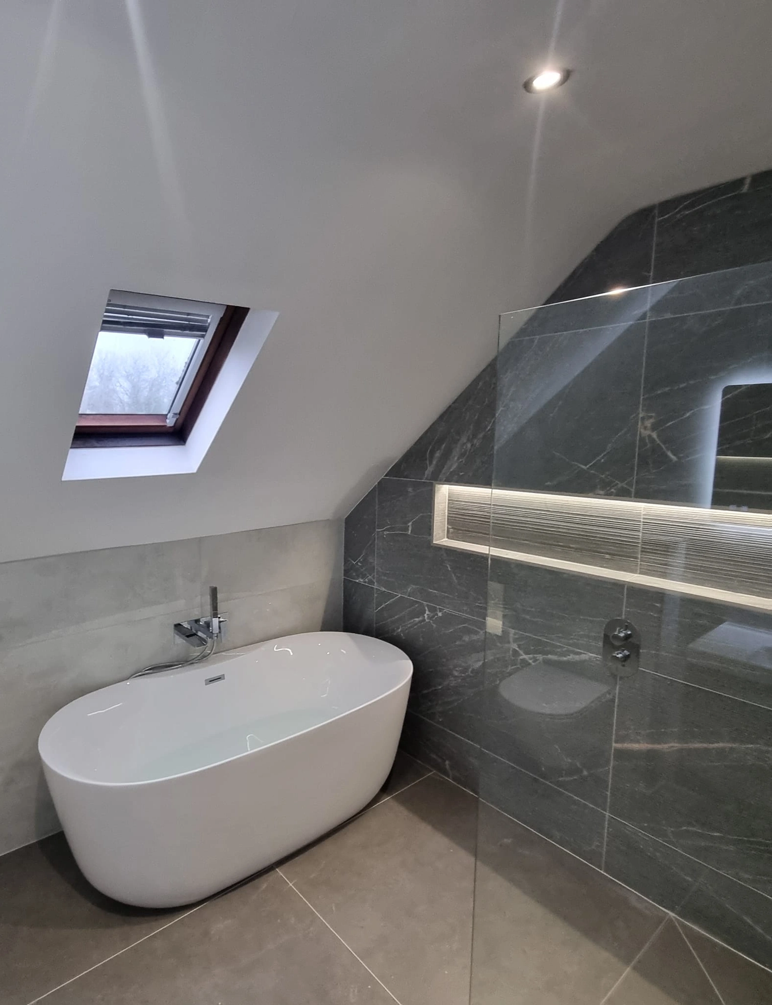 Modern bathroom with a white freestanding bathtub under a slanted ceiling with a skylight window and dark gray tiled walls.