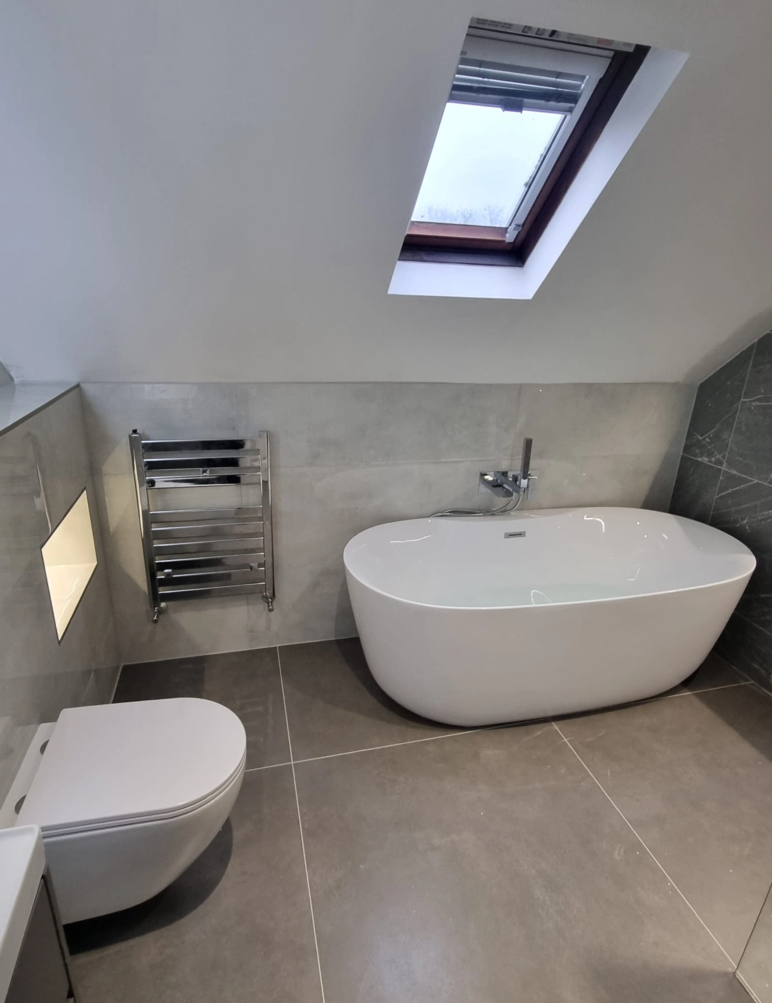 Modern bathroom with a white freestanding bathtub, wall-mounted toilet, chrome towel radiator, and skylight window.