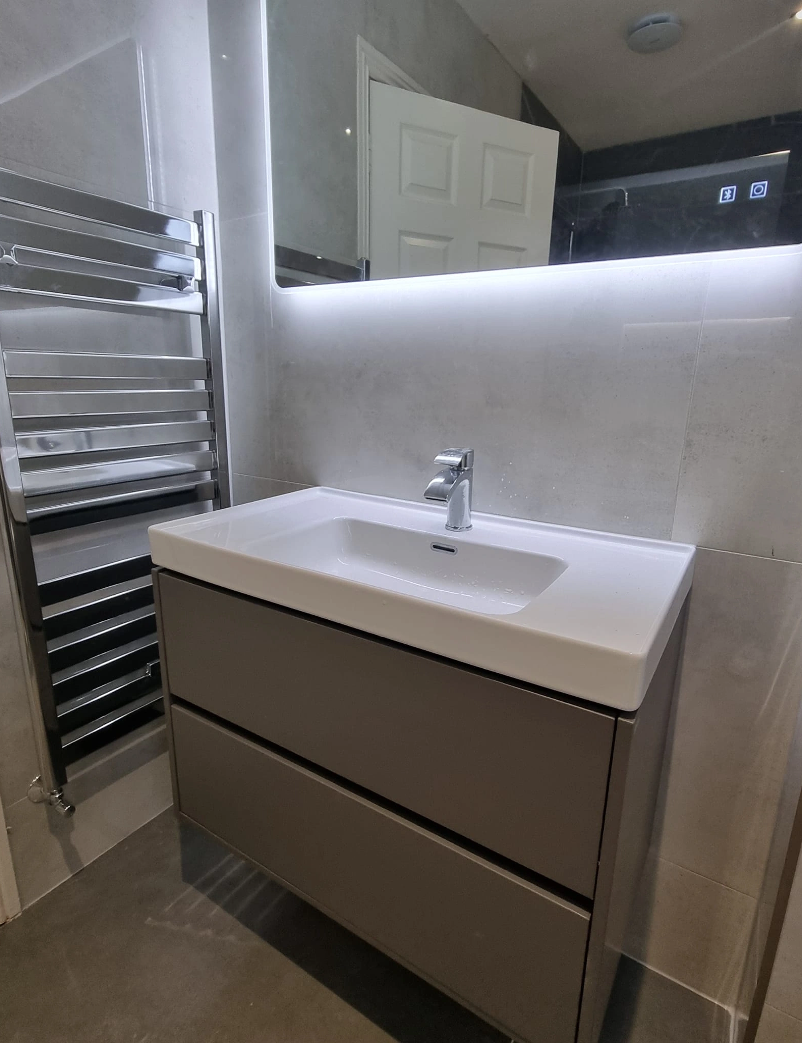 Modern bathroom sink with grey cabinet, chrome faucet, illuminated mirror, and stainless steel towel warmer.