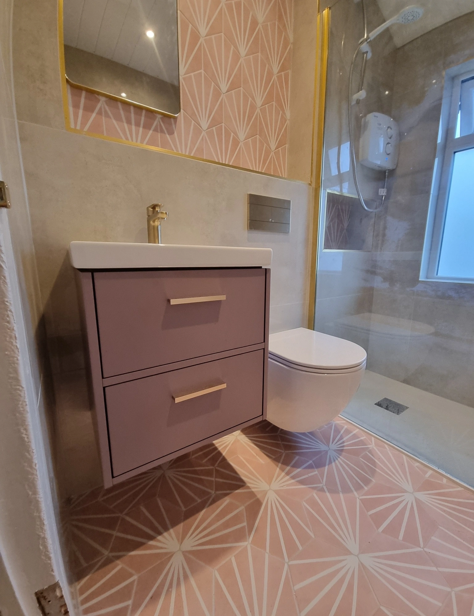 Modern bathroom with pink geometric patterned floor and wall tiles, a wall-mounted toilet, a purple vanity with a white sink, and a glass shower enclosure with a rainfall showerhead.