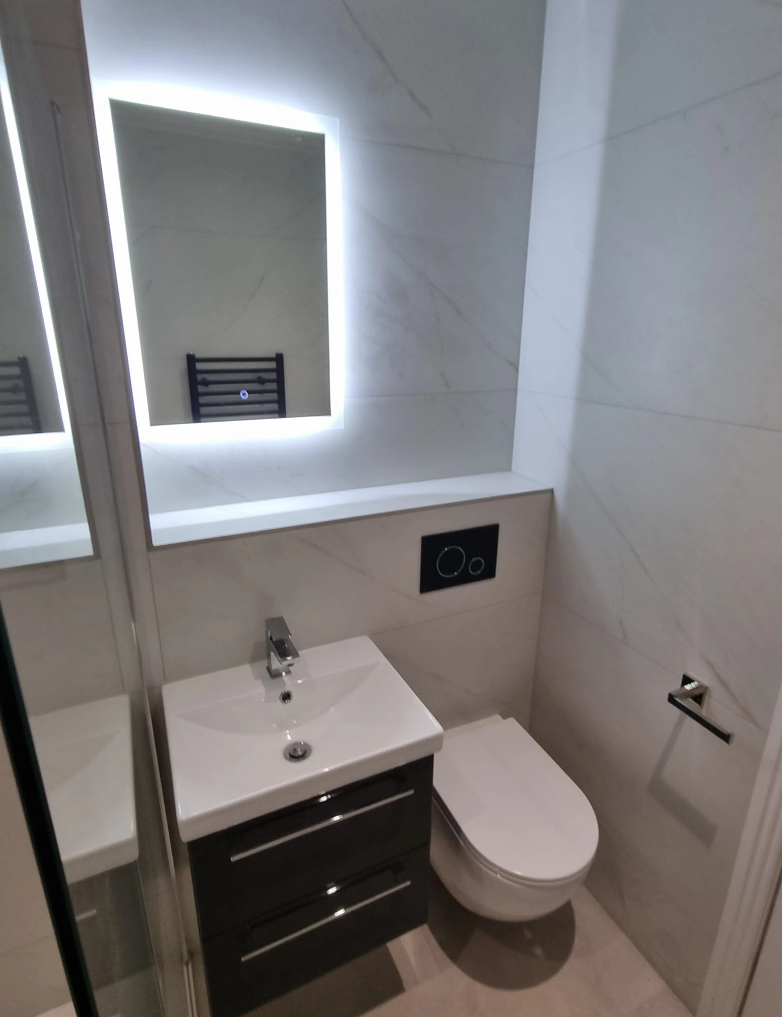 Modern small bathroom with a wall-mounted toilet, a white sink with a dark cabinet, and a backlit mirror.