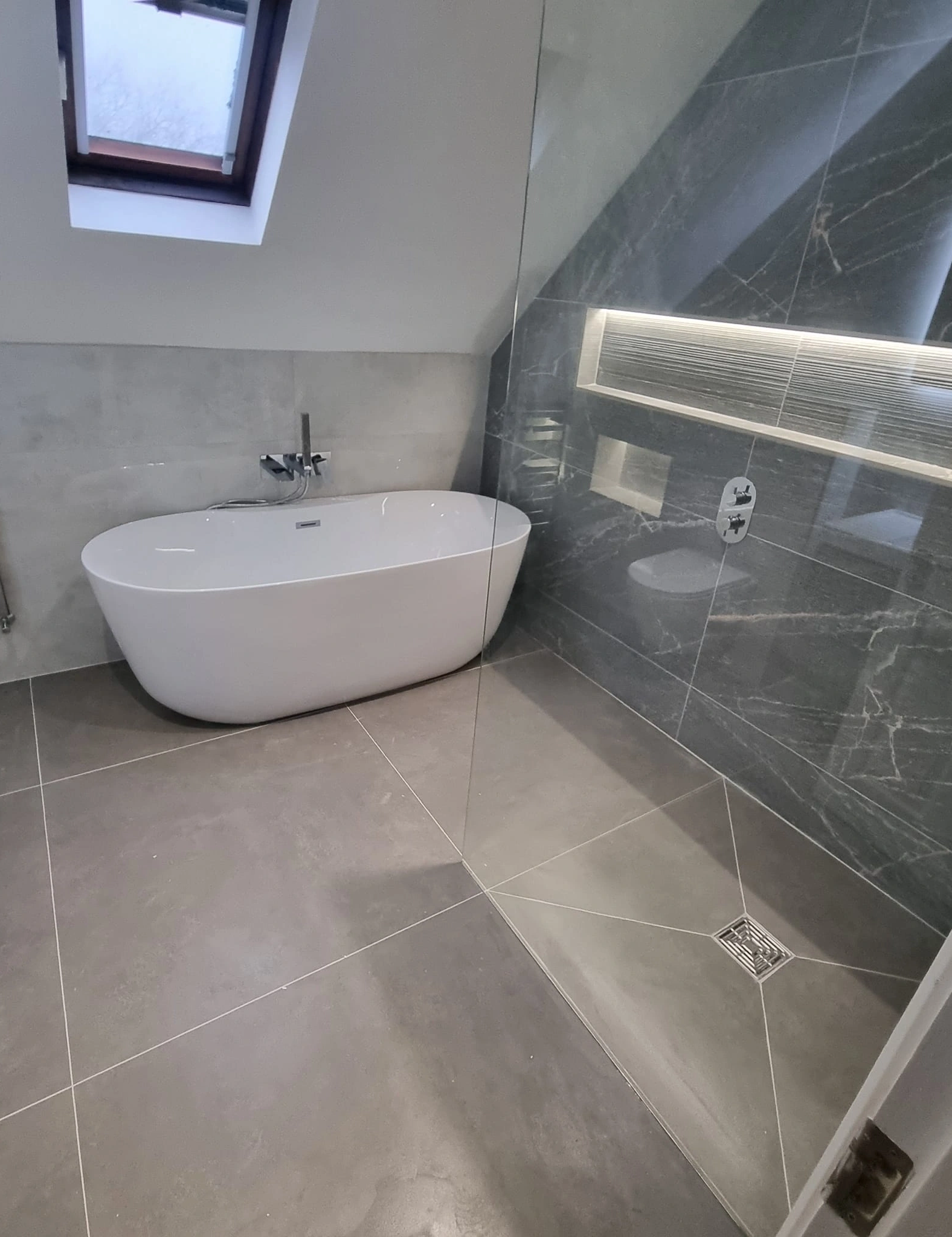 Modern bathroom with freestanding white bathtub under a skylight and a glass-enclosed shower with dark marble tiles.