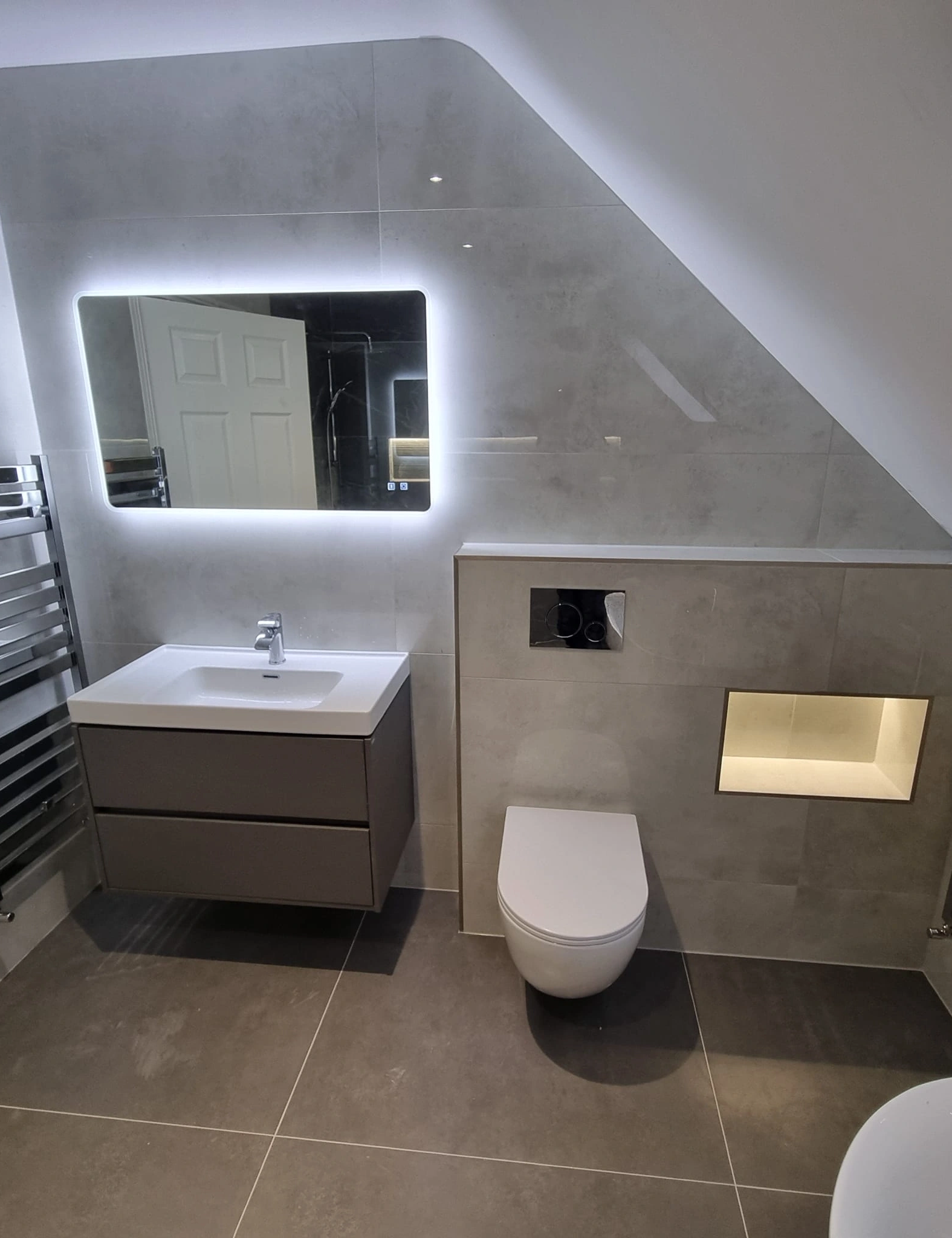 Modern bathroom with wall-mounted sink and cabinet, illuminated rectangular mirror, wall-mounted toilet, and built-in illuminated shelf.