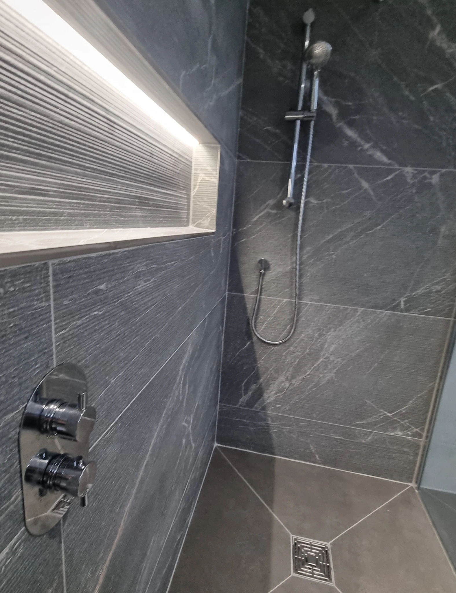 Modern shower with dark stone tiles, chrome fixtures, illuminated recessed shelf, and square floor drain.