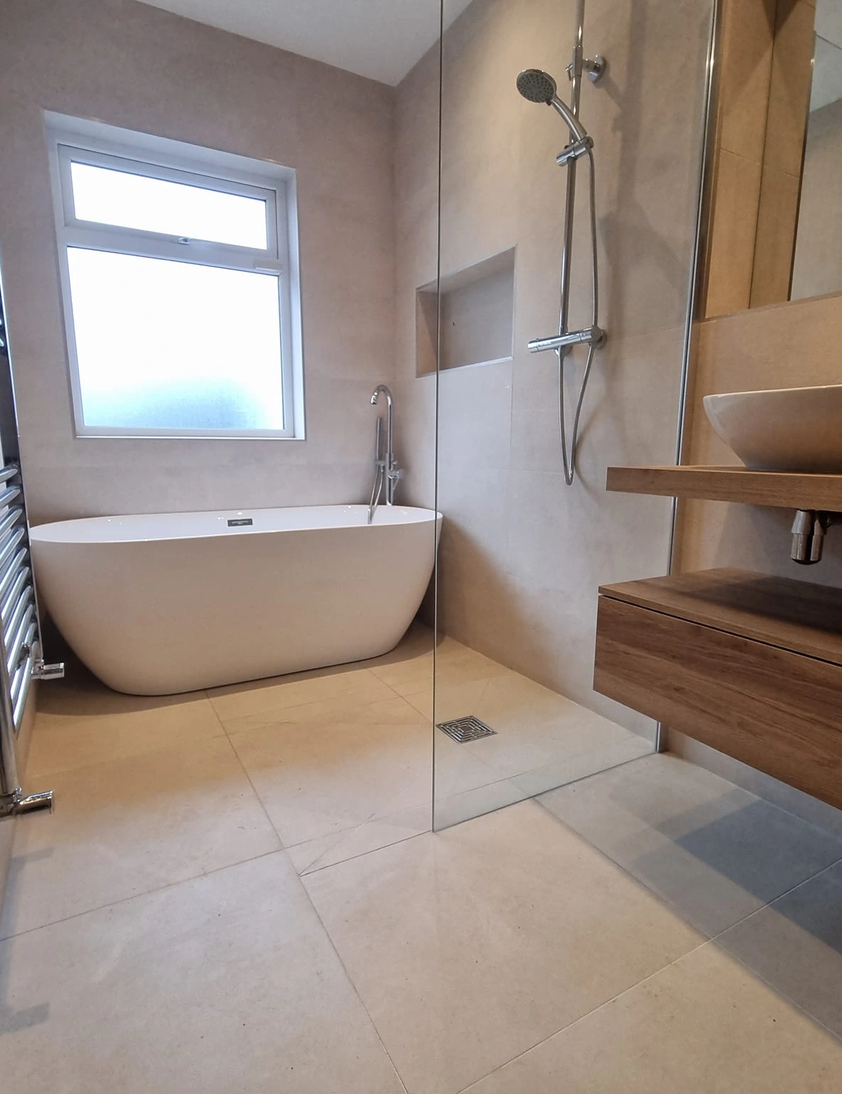 Modern bathroom with a white freestanding bathtub, walk-in glass shower, wooden vanity with vessel sink, and large frosted window.