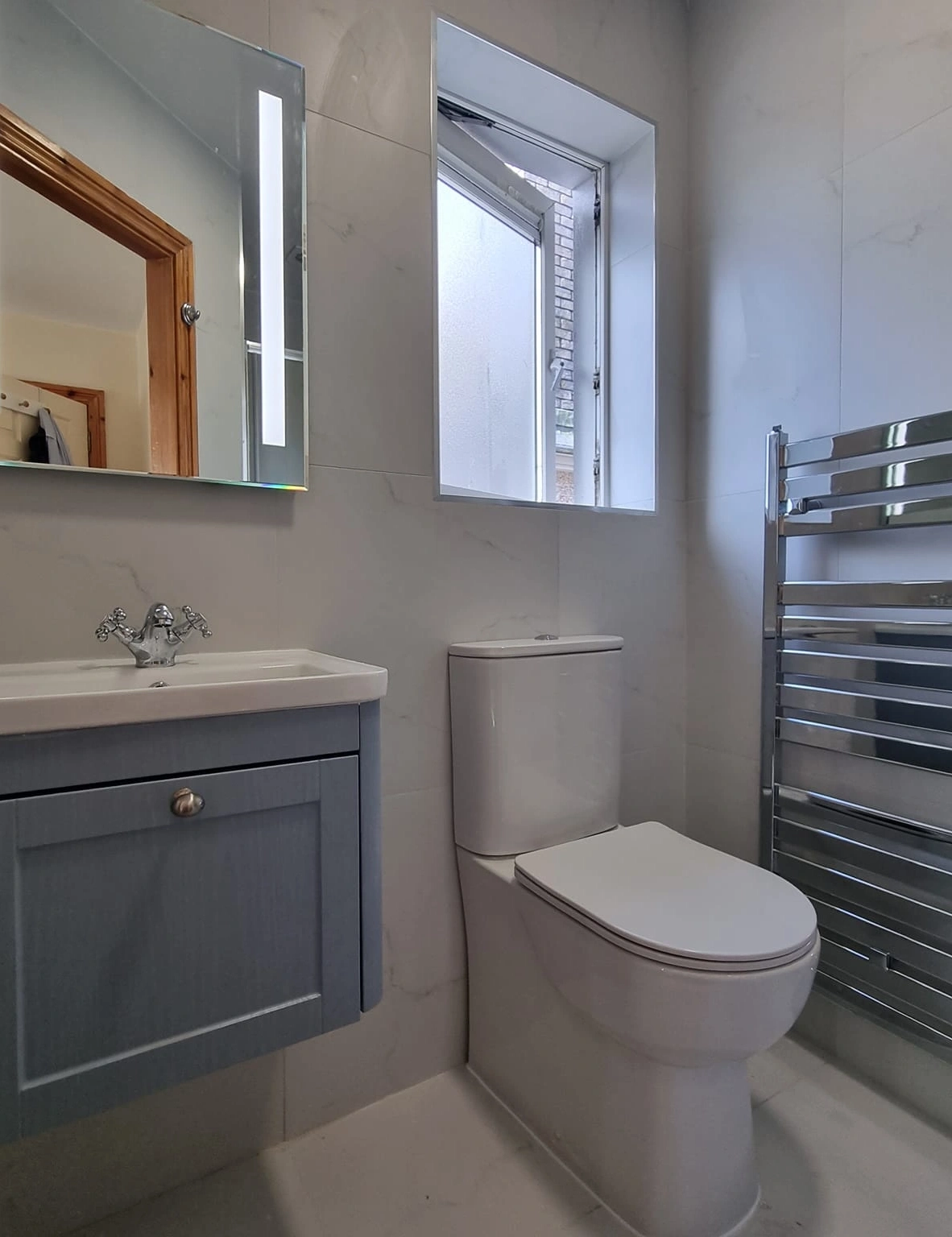 Modern bathroom with a white toilet, gray vanity with sink, rectangular mirror with light strip, open frosted window, and chrome towel radiator.