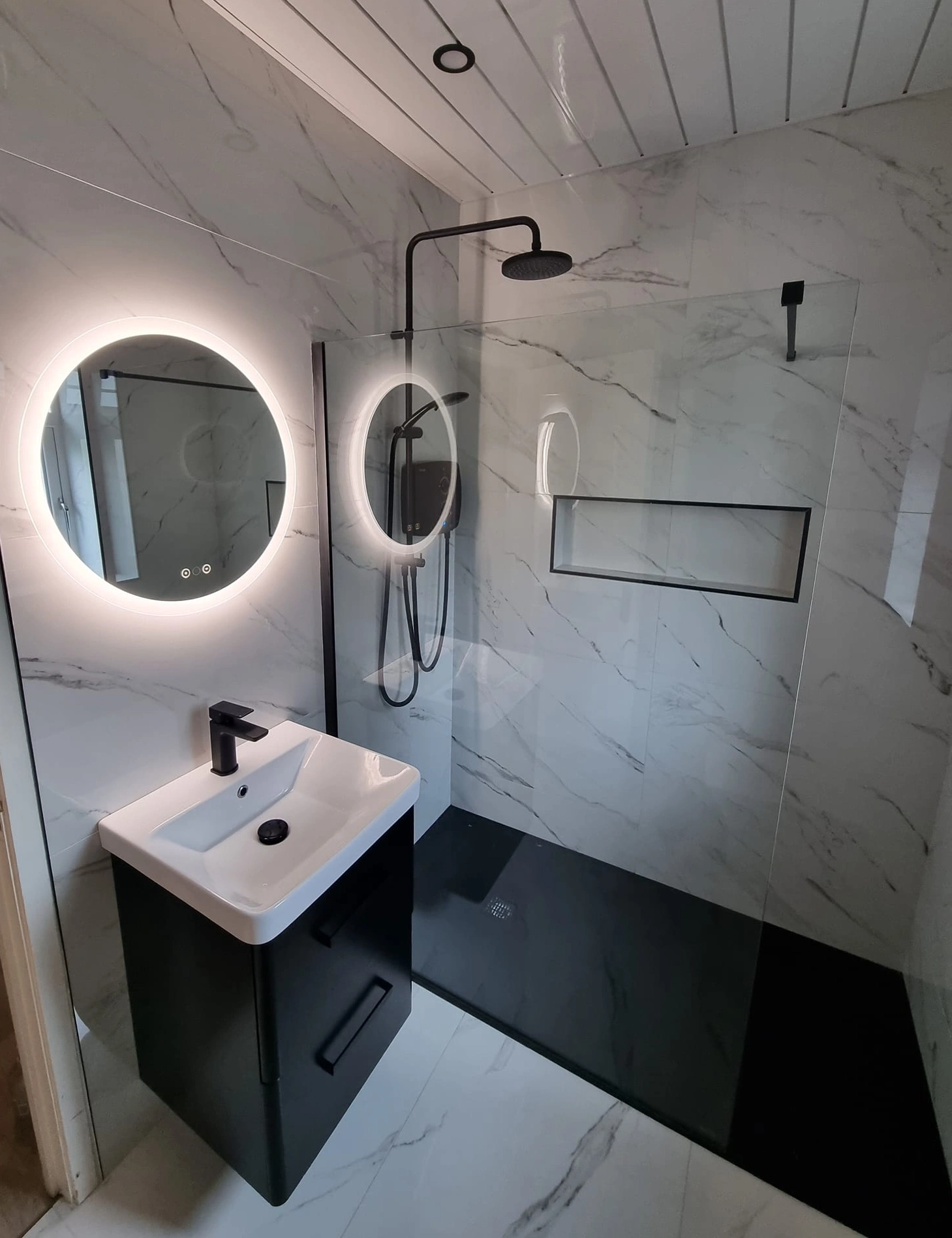 Modern bathroom with marble walls, a round backlit mirror above a black vanity with white sink, and a black shower area with a glass partition.