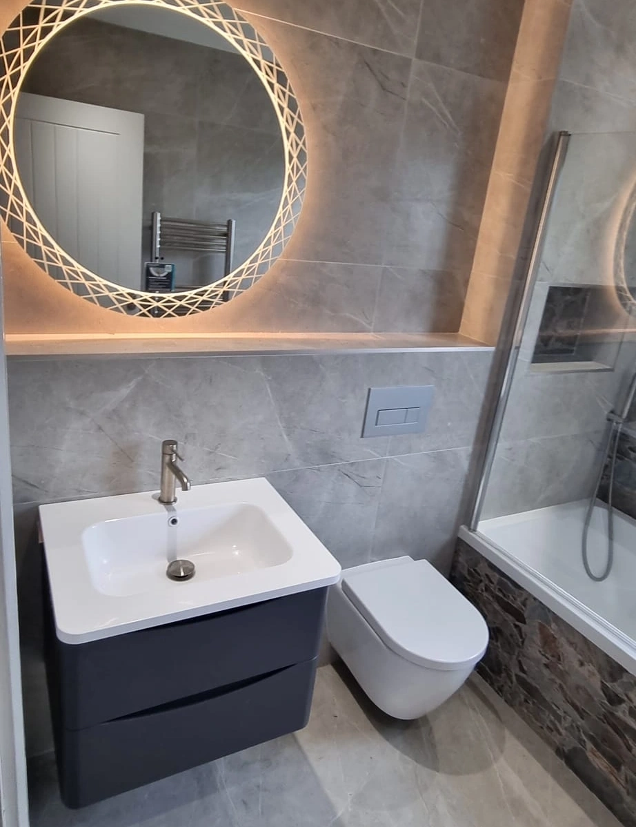 Modern bathroom with a wall-mounted toilet, gray vanity with white sink, round illuminated mirror, and a bathtub with glass screen.