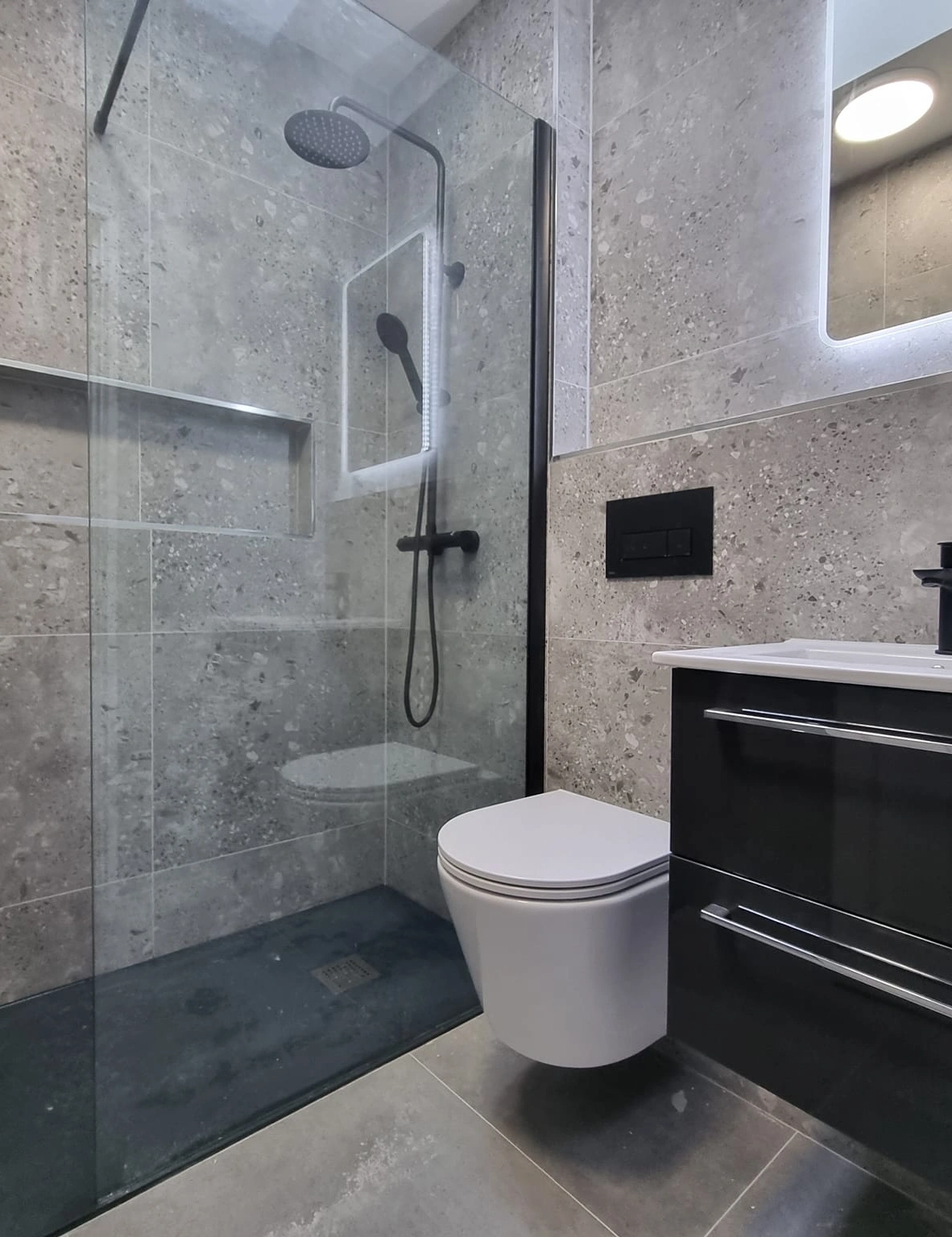 Modern bathroom with a glass-enclosed shower featuring a black showerhead, a wall-mounted white toilet, and a black vanity with a white sink.
