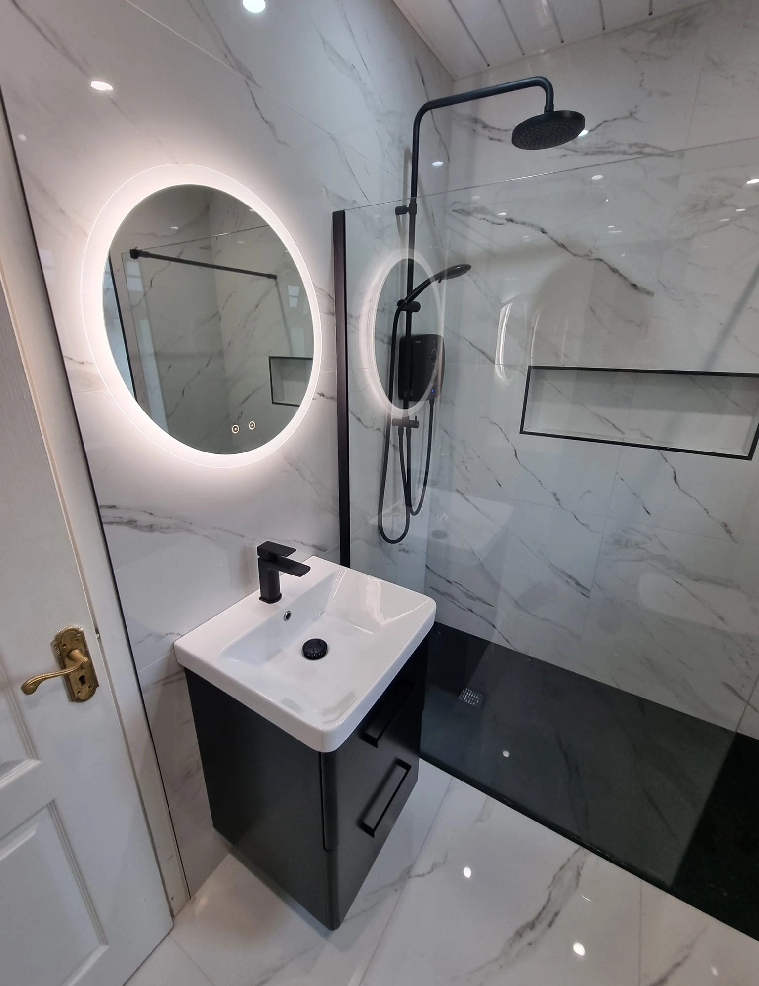 Modern bathroom with black and white color scheme featuring a black vanity with white sink, a round illuminated mirror, and a walk-in shower with black fixtures and glass partition.