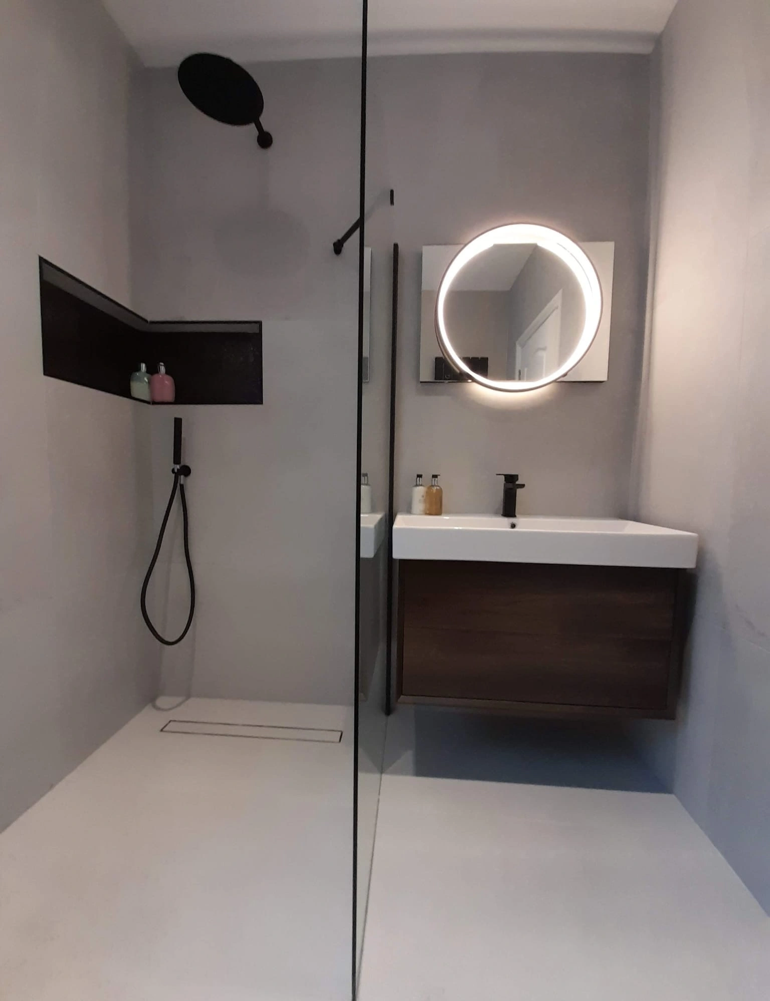 Minimalist bathroom with a walk-in shower featuring a black showerhead and hose, a rectangular niche with two bottles, and a floating sink cabinet under a round illuminated mirror.