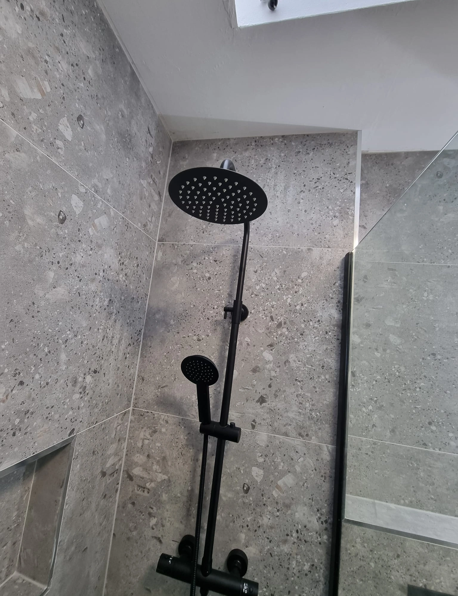 Modern black dual showerhead system mounted on a gray tiled bathroom wall with a glass partition.