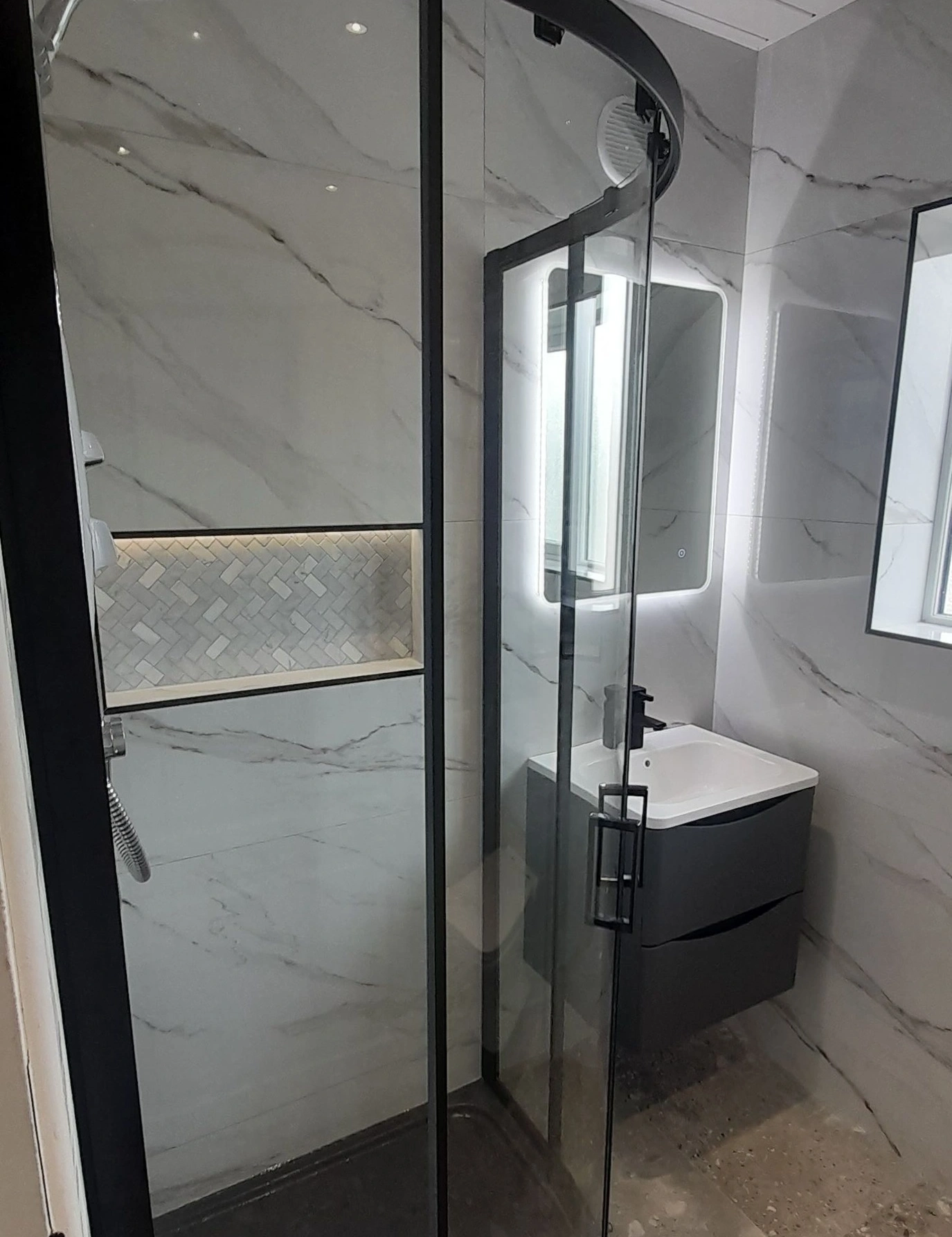 Compact modern bathroom with glass shower enclosure, white marble walls, and a wall-mounted sink with illuminated mirror.