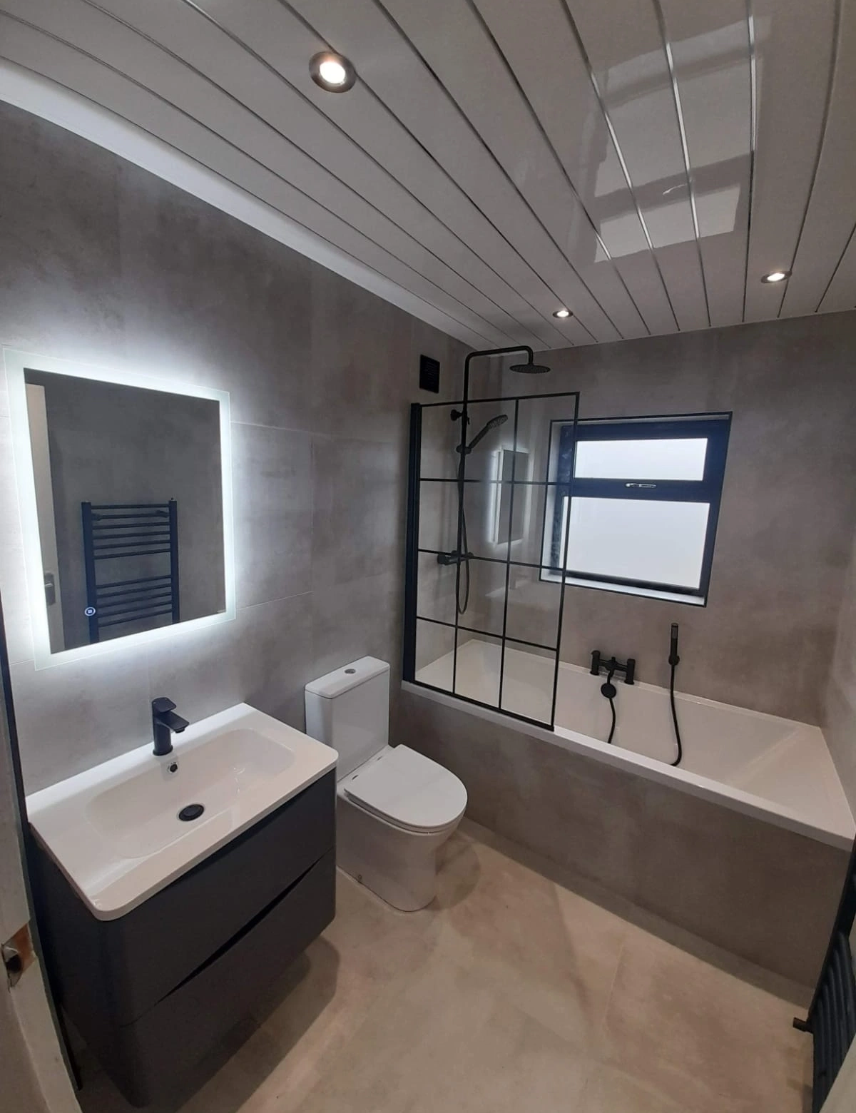Modern bathroom with gray tiled walls and floor, a white toilet, black vanity with white sink, illuminated mirror, bathtub with black shower fixtures, and a window.
