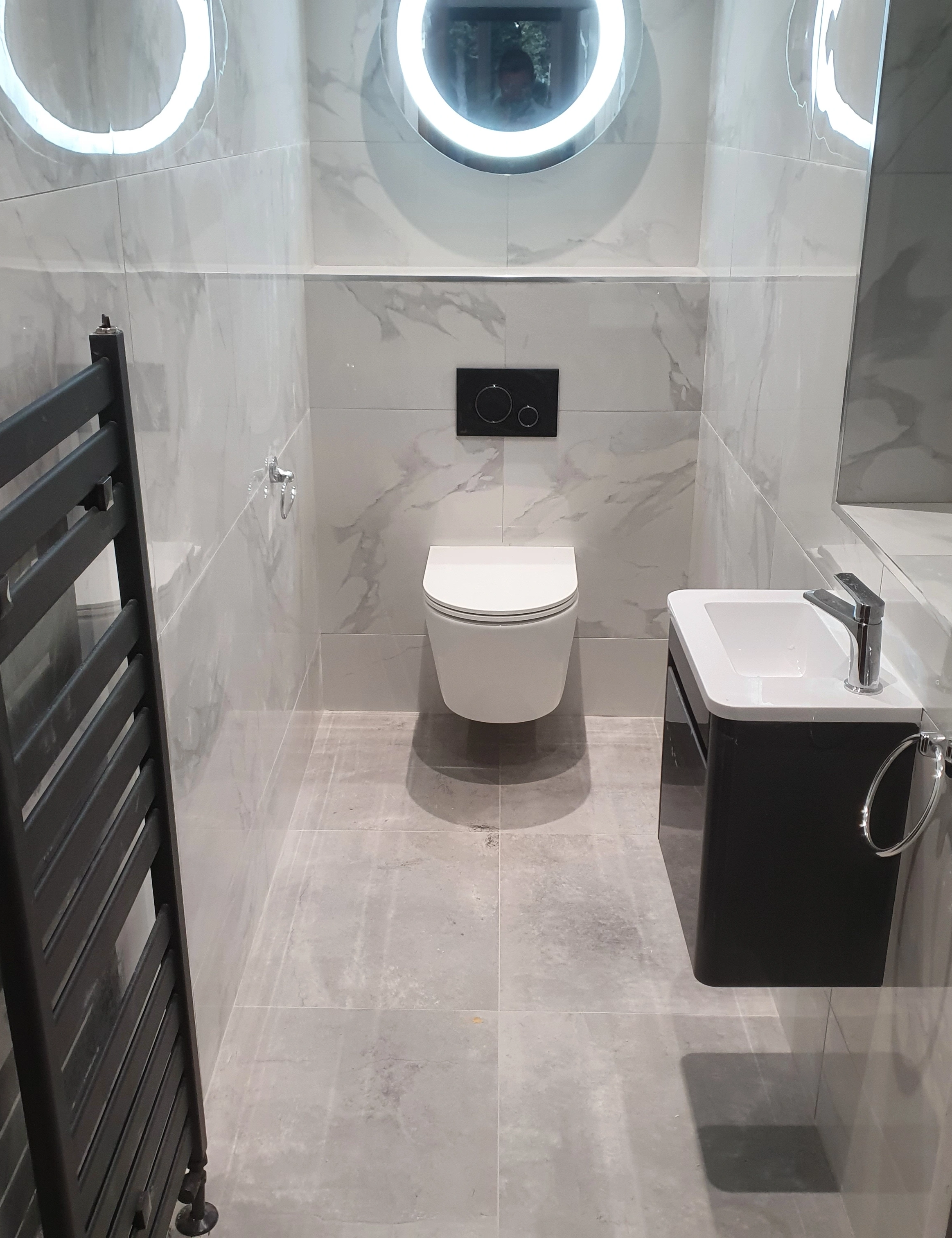 Modern small bathroom with wall-mounted toilet, black towel radiator, and compact sink with cabinet and mirror.