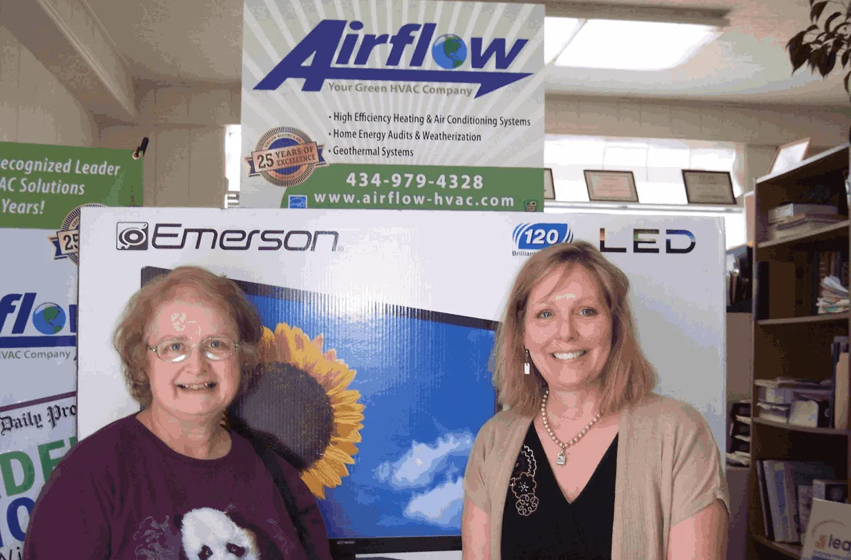 Two smiling women standing in front of an Emerson LED TV box inside an office with Airflow HVAC company banners.