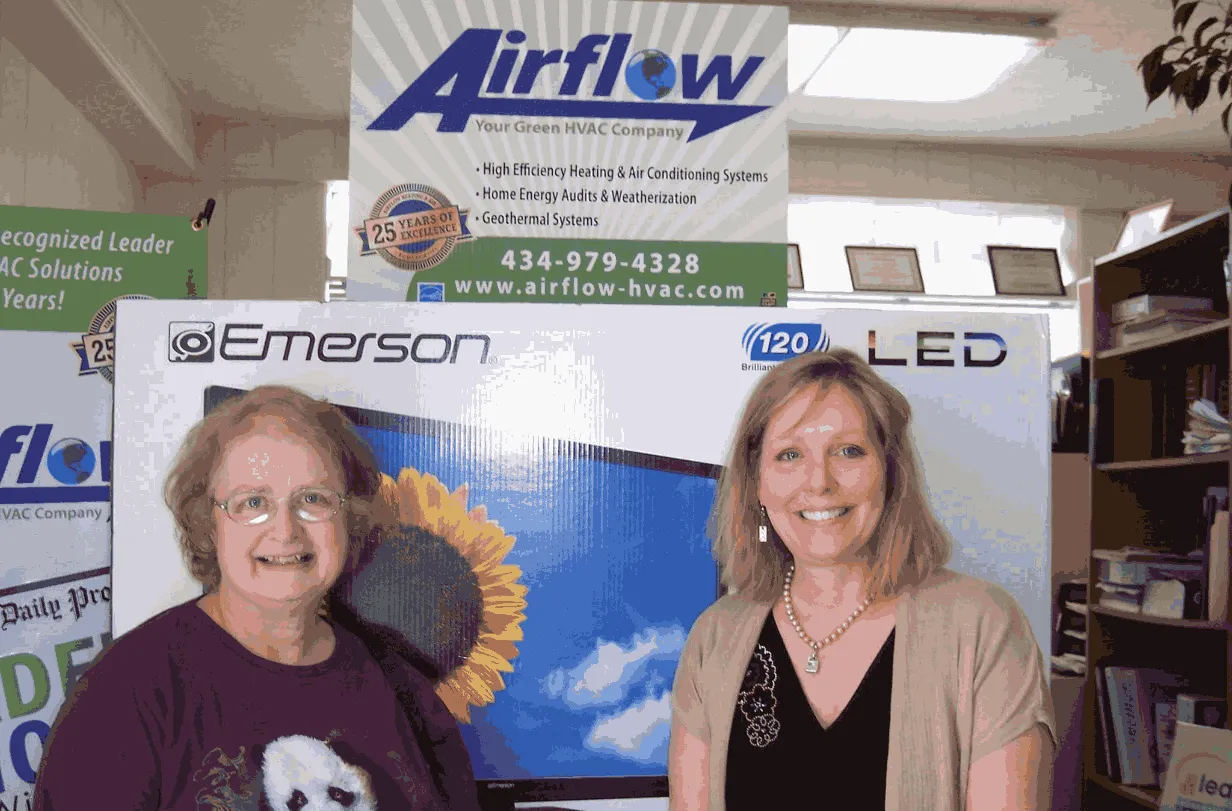 Two women smiling and standing in front of Emerson LED TV box and Airflow HVAC company signage indoors.