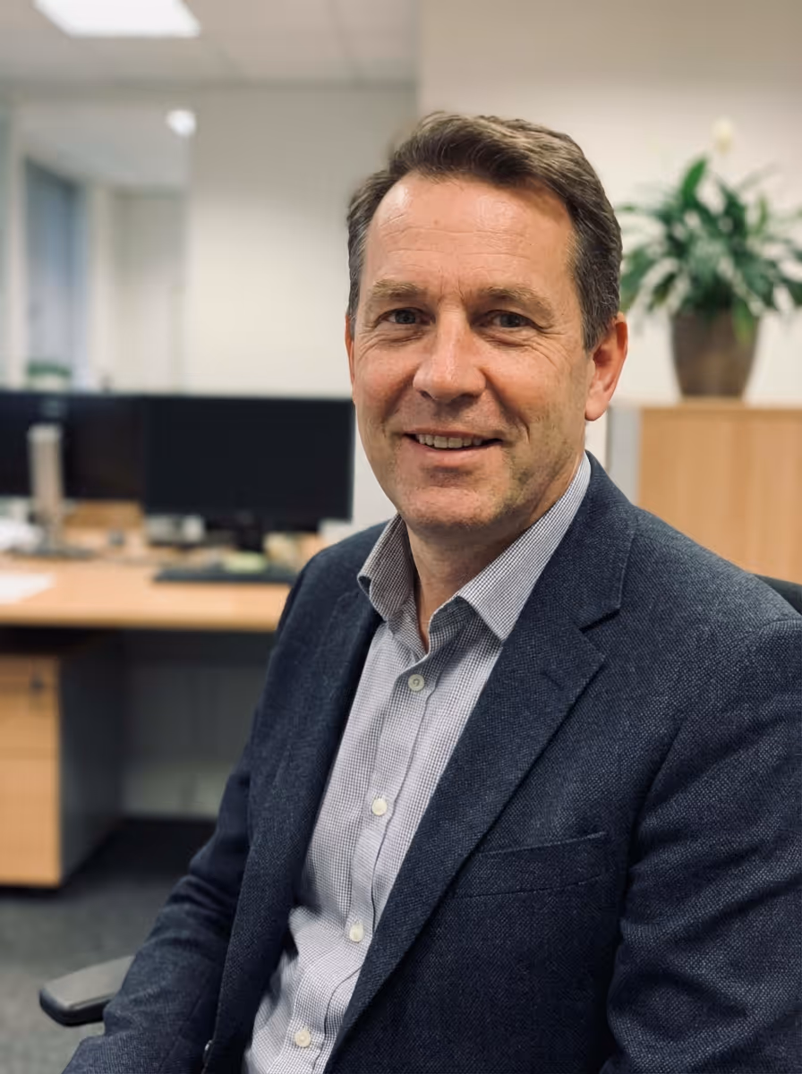 Middle-aged man in a gray blazer and light checkered shirt smiling in an office setting.
