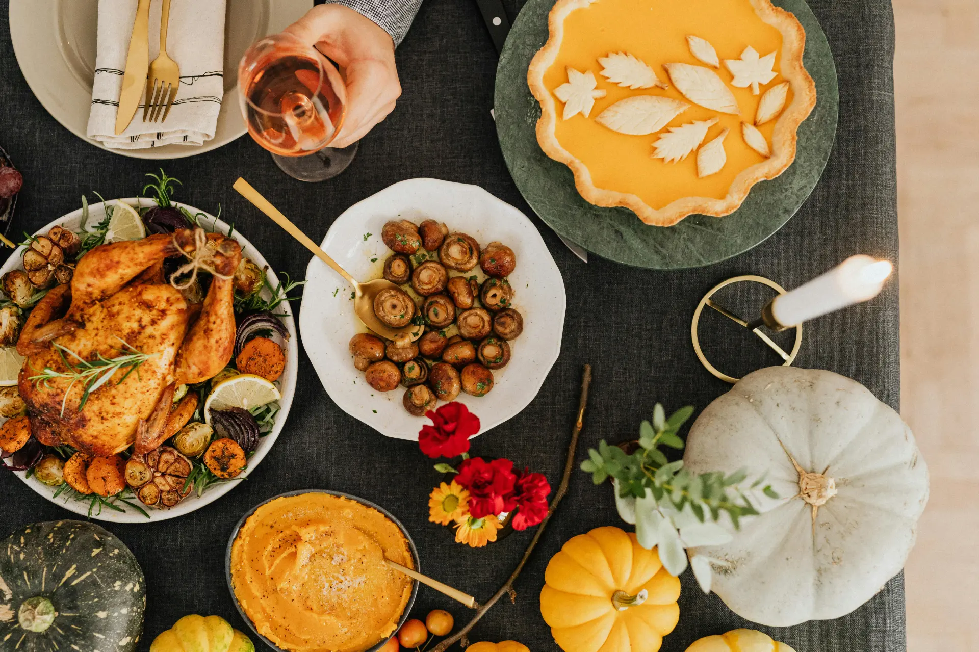 A festive table setting featuring a roasted chicken, pumpkin pie with leaf designs, a bowl of sautéed mushrooms, candles, and autumn decor. A hand holds a glass of wine, adding a celebratory tone.