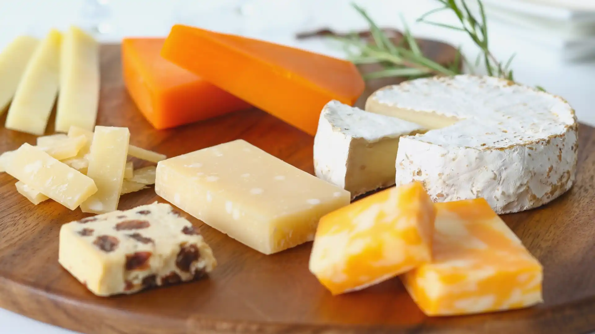 A wooden platter adorned with assorted cheeses: creamy brie, vibrant orange cheddar, marble, and more. Fresh rosemary adds an elegant touch.