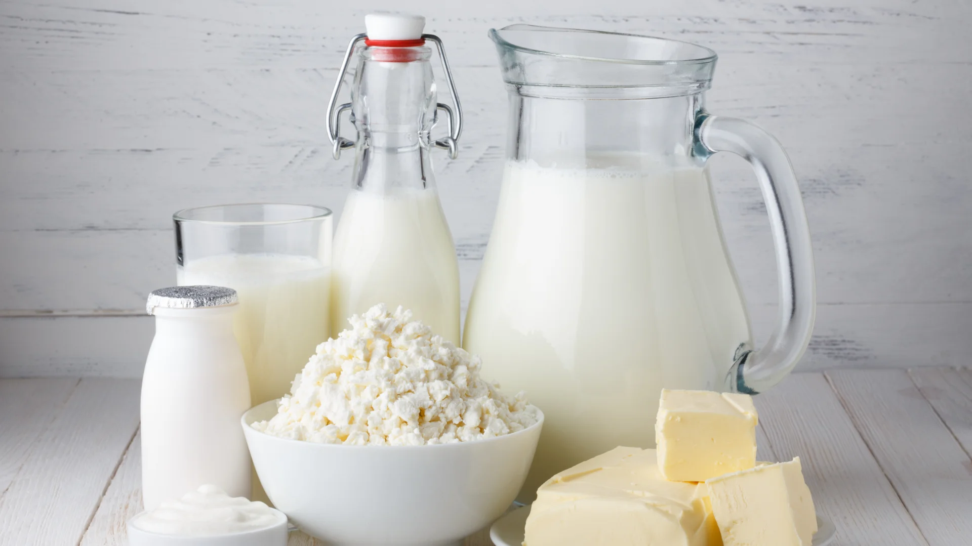 Assorted dairy products on a wooden table include a glass jug and bottle of milk, a small bottle of yogurt, butter blocks, and a bowl of cottage cheese. Creamy and fresh tone.