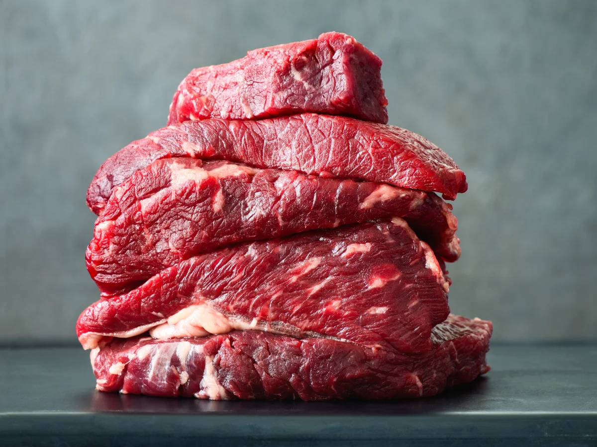 A stack of raw red meat cuts, layered on a dark surface against a textured gray background. The fresh meat appears marbled and ready for cooking.
