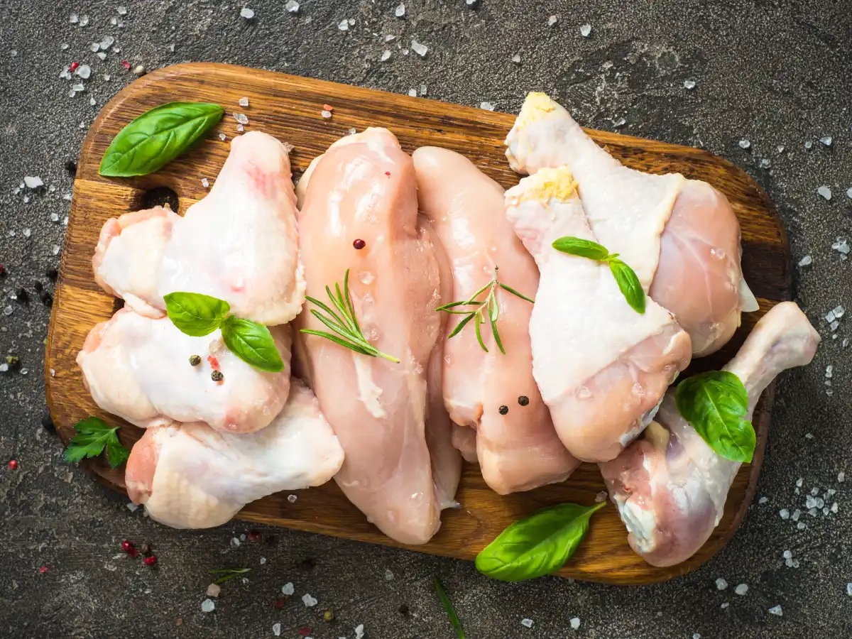 Raw chicken pieces on a wooden cutting board, garnished with basil leaves and rosemary. Salt and peppercorns are scattered around, creating a fresh, rustic feel.