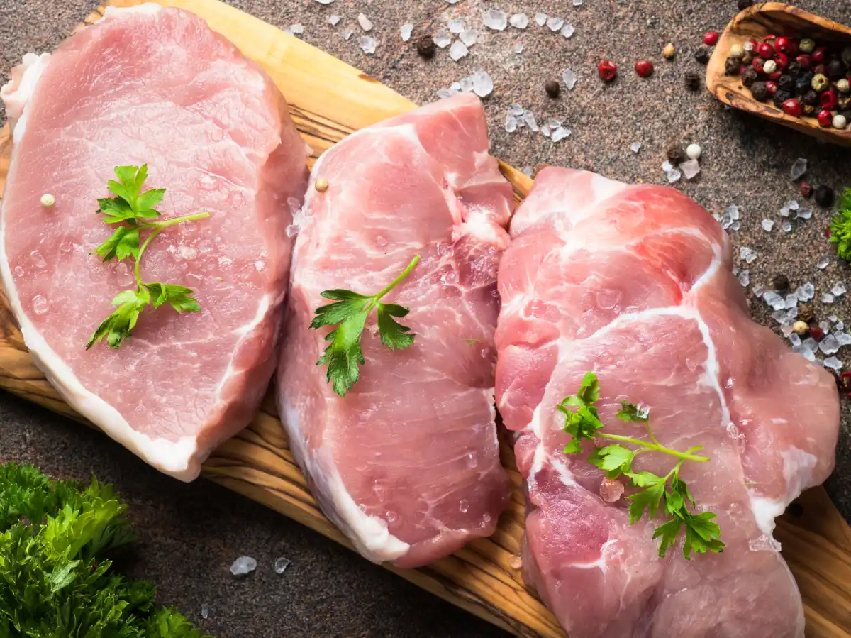 Three raw pork chops topped with parsley on a wooden cutting board. Surrounding are scattered peppercorns, coarse salt, and fresh herbs.