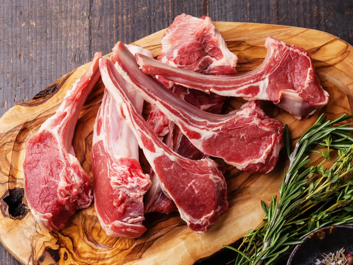 Raw lamb chops on a wooden board with sprigs of fresh rosemary beside them. The setting suggests a rustic, hearty cooking vibe.