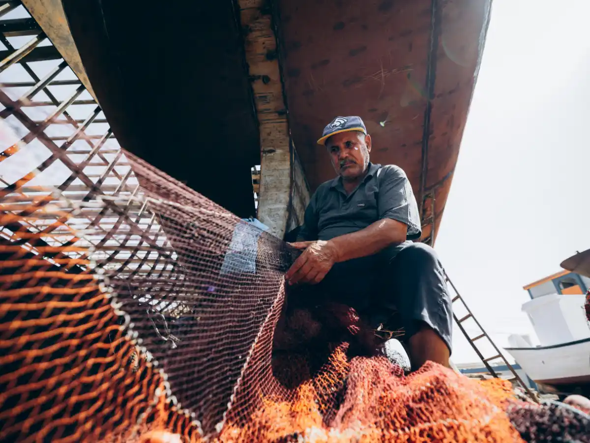 Elderly man repairs fishing net under a weathered boat. Sunlight filters through, highlighting his focused expression. Rustic, maritime setting.