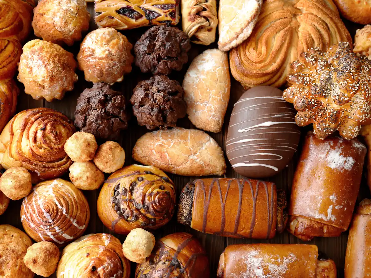 Assorted pastries including croissants, chocolate chip cookies, muffins, and bread rolls are arranged in a visually appealing, colorful pattern.