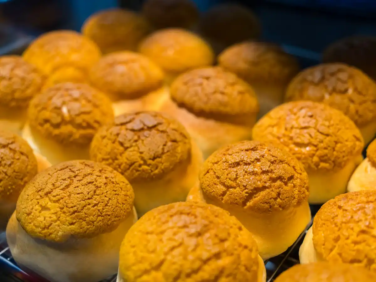 Golden-brown pineapple buns with a crispy, cracked top are closely arranged on a baking tray. The image conveys warmth and freshness.