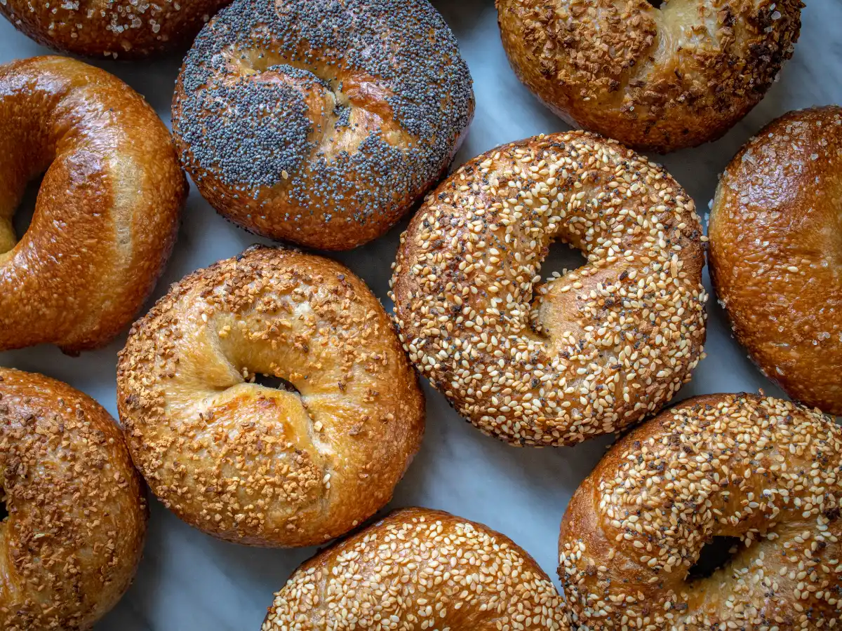 A variety of bagels arranged on a marble surface, featuring toppings like poppy seeds, sesame seeds, and toasted onions. The image conveys a fresh, appetizing feel.